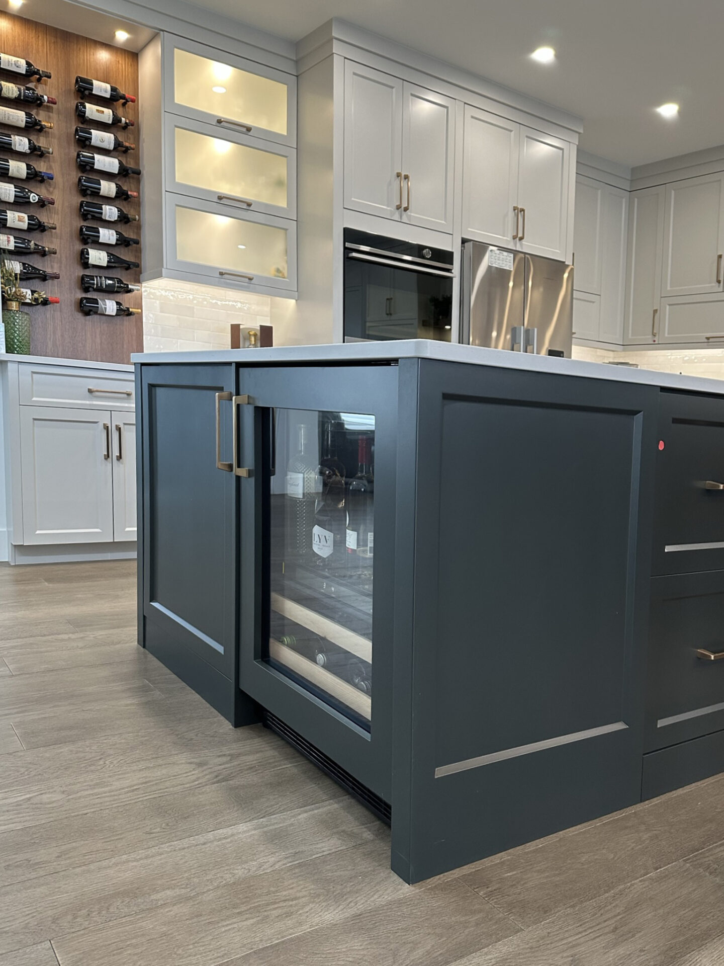 A modern kitchen with a gray island featuring a wine cooler, white cabinetry, a wine rack with bottles, and stainless steel appliances.