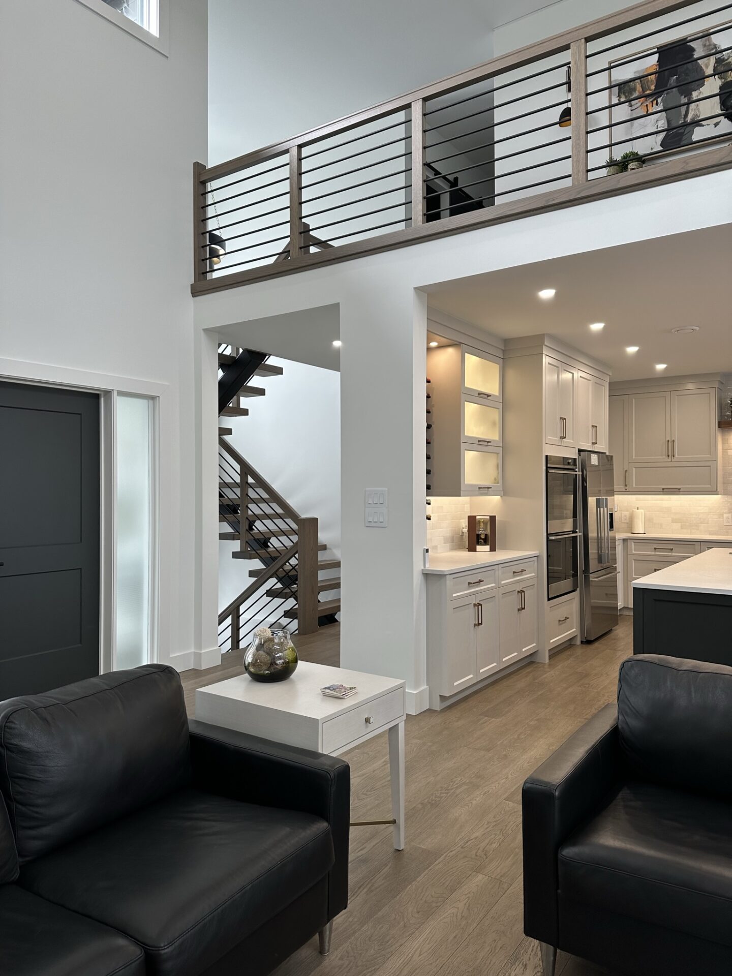 A modern interior with a sitting area, staircase, upper-level hallway with railing, and kitchen with white cabinets and stainless steel appliances.