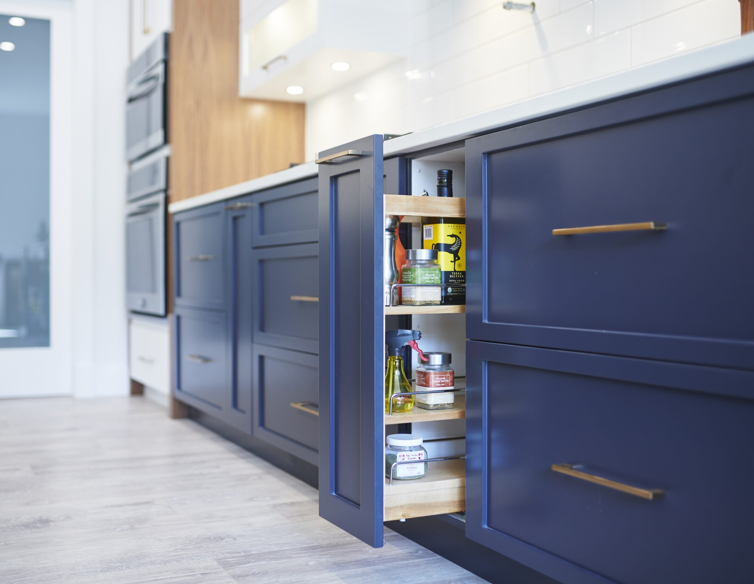 A modern kitchen with blue cabinets featuring brass handles. An open pull-out drawer reveals organized bottles and condiments. Bright, clean, and contemporary design.