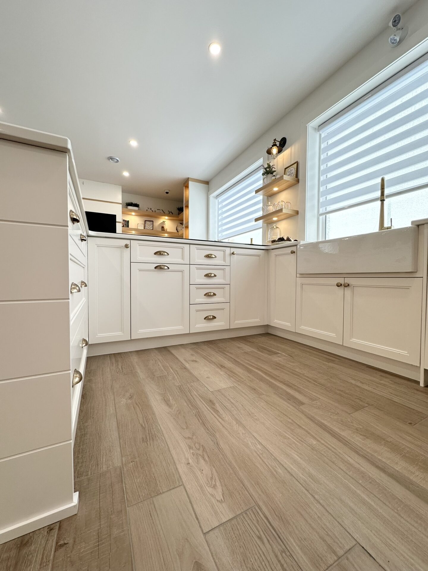 Bright kitchen interior with white cabinets, drawers with gold handles, wood flooring, and natural light coming through a window with blinds.