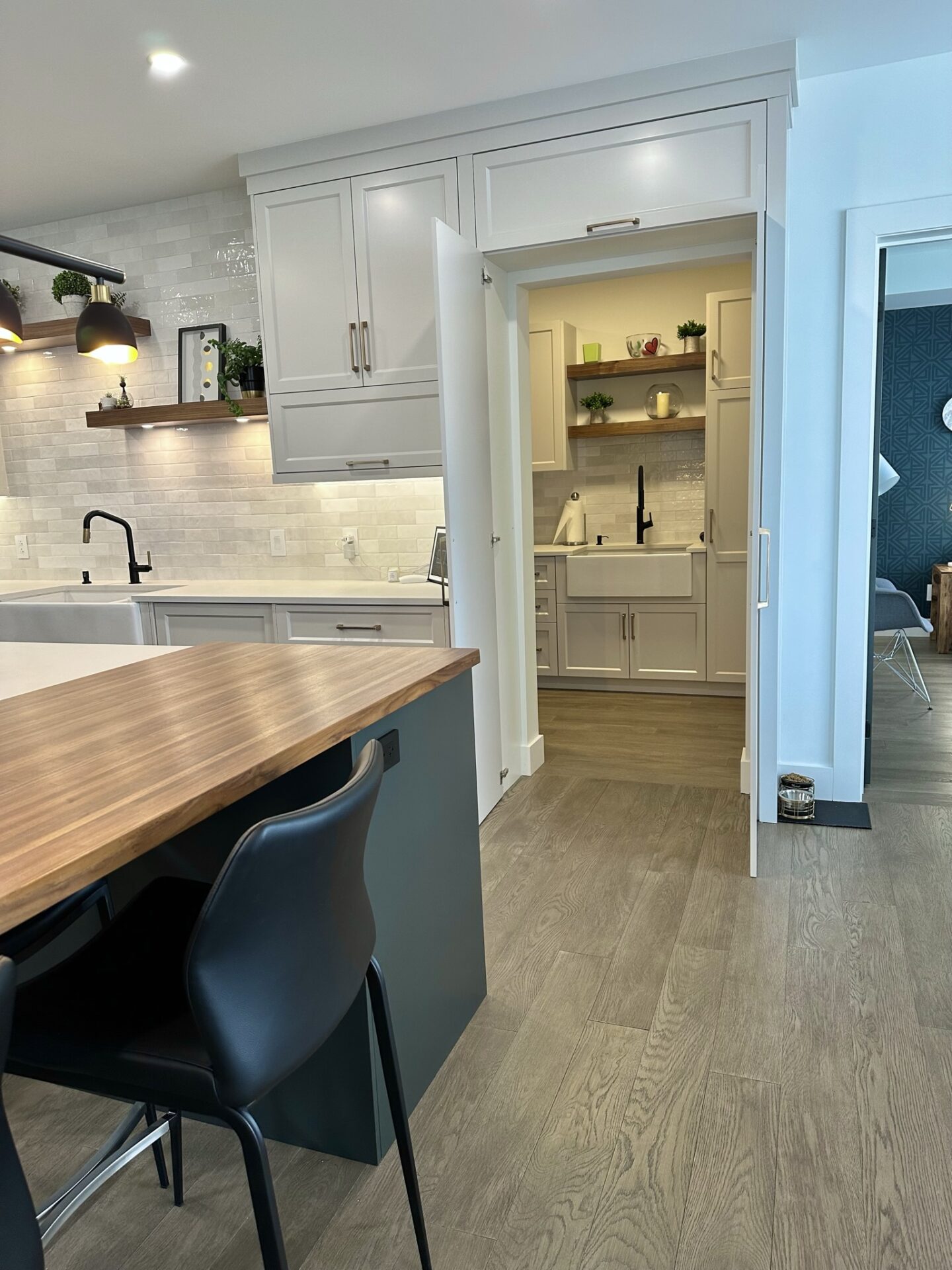 The image shows a modern kitchen with white cabinets, a wooden countertop, black chairs, stainless steel appliances, and a herringbone tile backsplash.