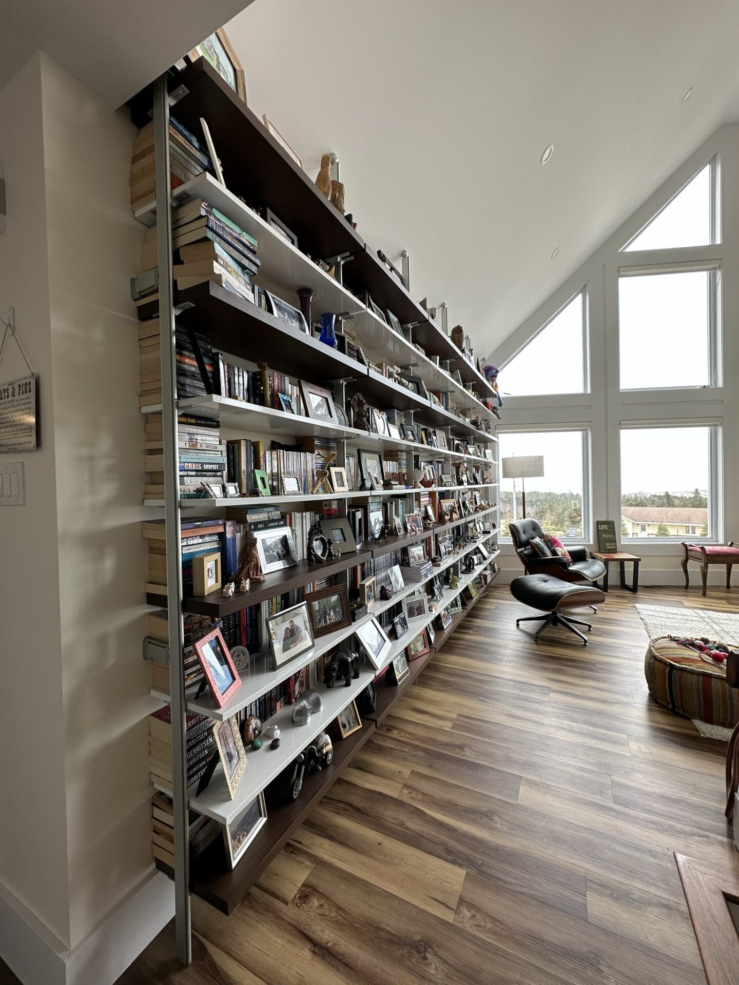 A spacious room with a large bookshelf wall filled with books, photos, and objects, wooden flooring, large windows, and a modern lounge chair.