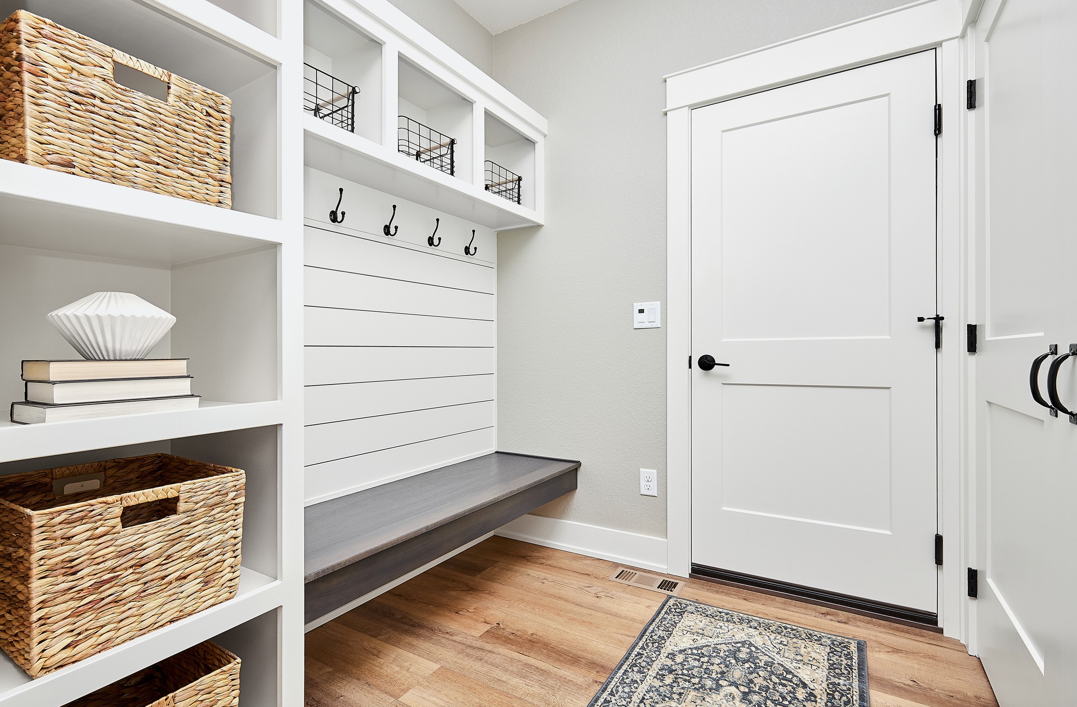 A modern, clean mudroom featuring white built-in shelves, a bench with hooks above, a closed white door, woven baskets, books, and a decorative rug.