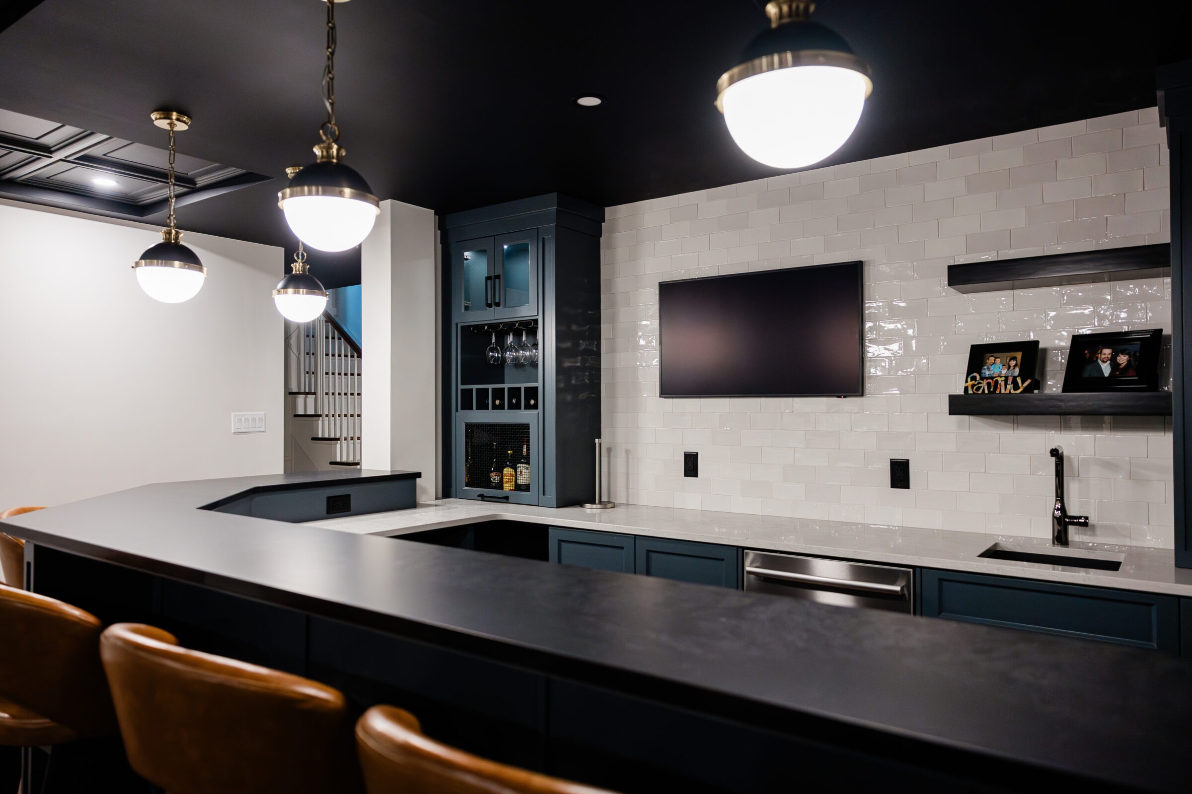 This image shows a stylish home bar area with dark cabinetry, a sink, elegant pendant lighting, a television, and subway tile backsplash.
