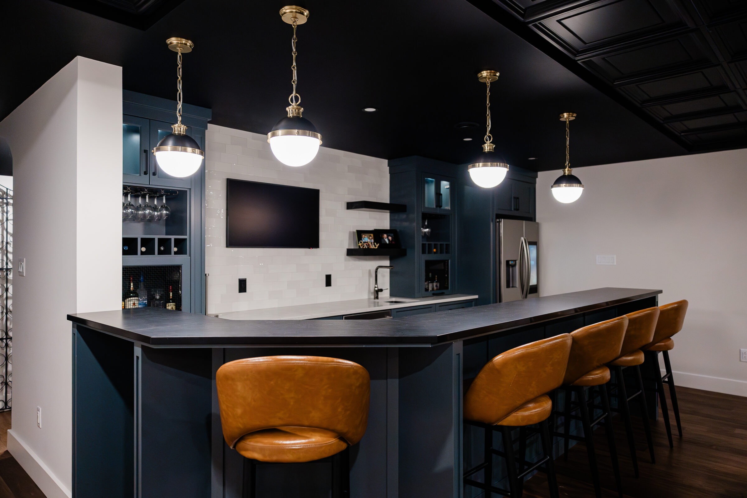 A modern home bar with dark interiors, pendant lighting, tan bar stools, glossy black countertop, and built-in cabinetry with wine storage.