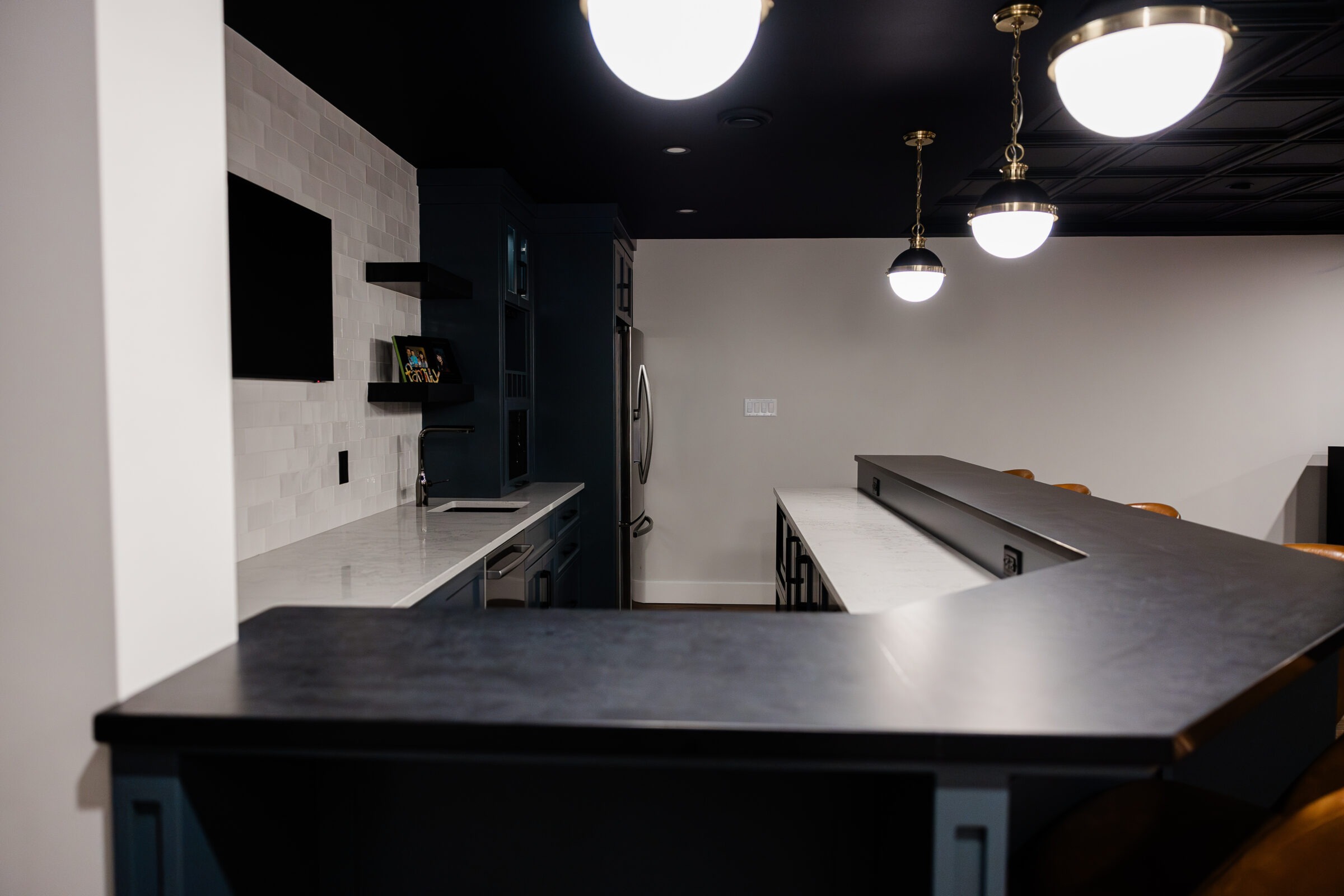 Modern kitchen interior with dark cabinets, white countertops, built-in appliances, pendant lights, and a bar with stools. The space is neat and uncluttered.