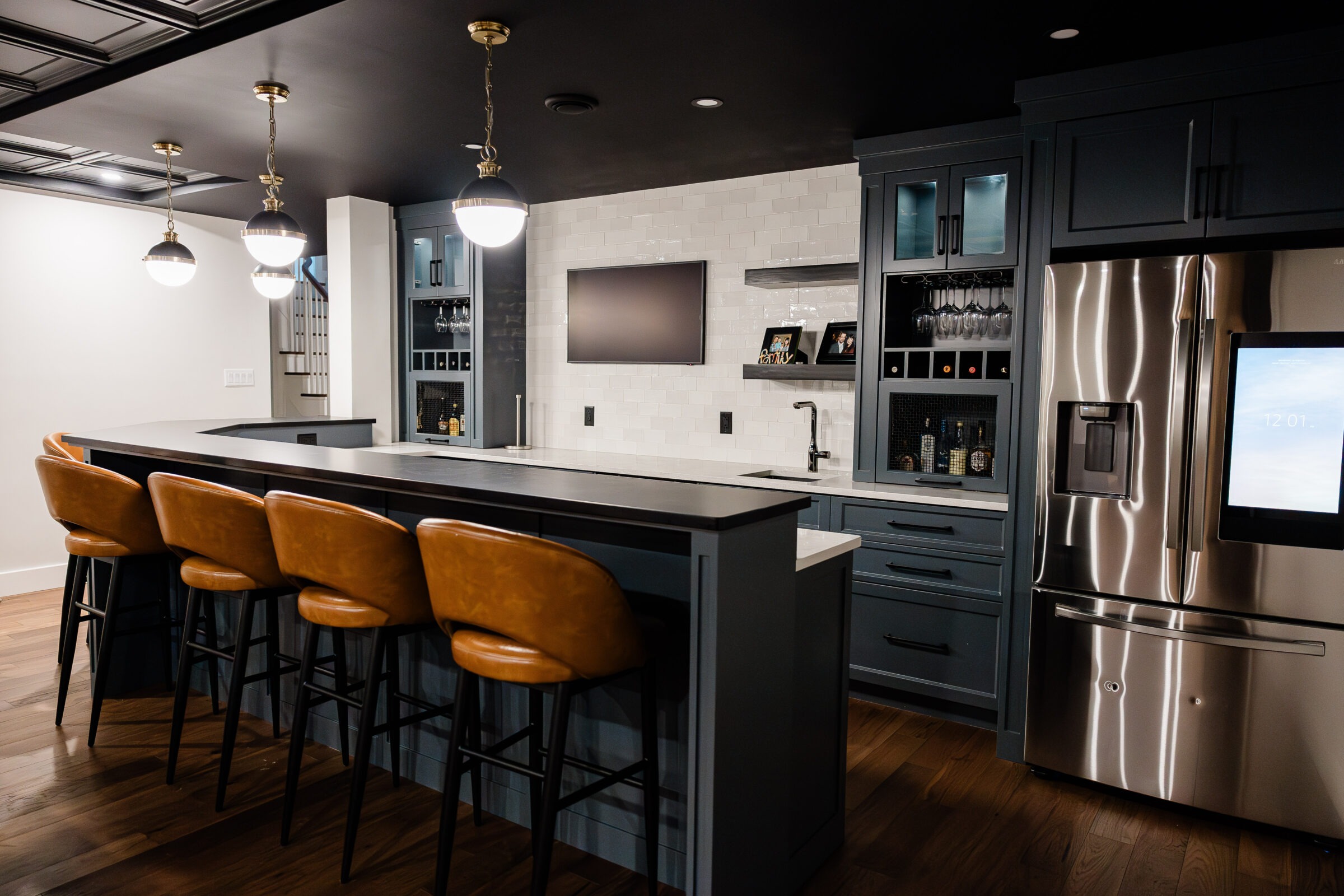 A modern kitchen with dark blue cabinets, stainless steel appliances, tan leather bar stools, and pendant lighting over a central island.