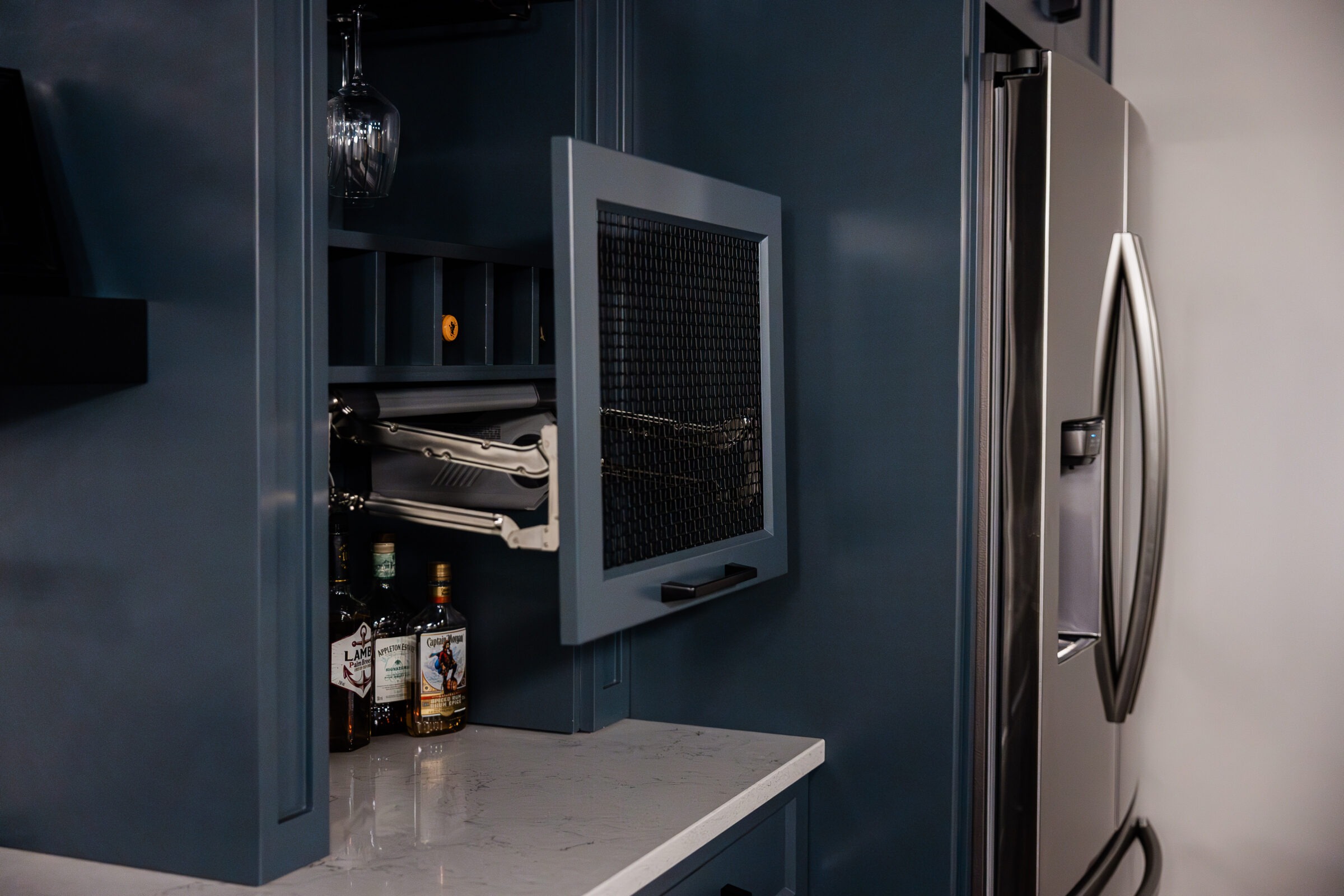 A modern kitchen cabinet with an open door, revealing a hidden rack with bottles of liquor and a hanging wine glass, next to a refrigerator.