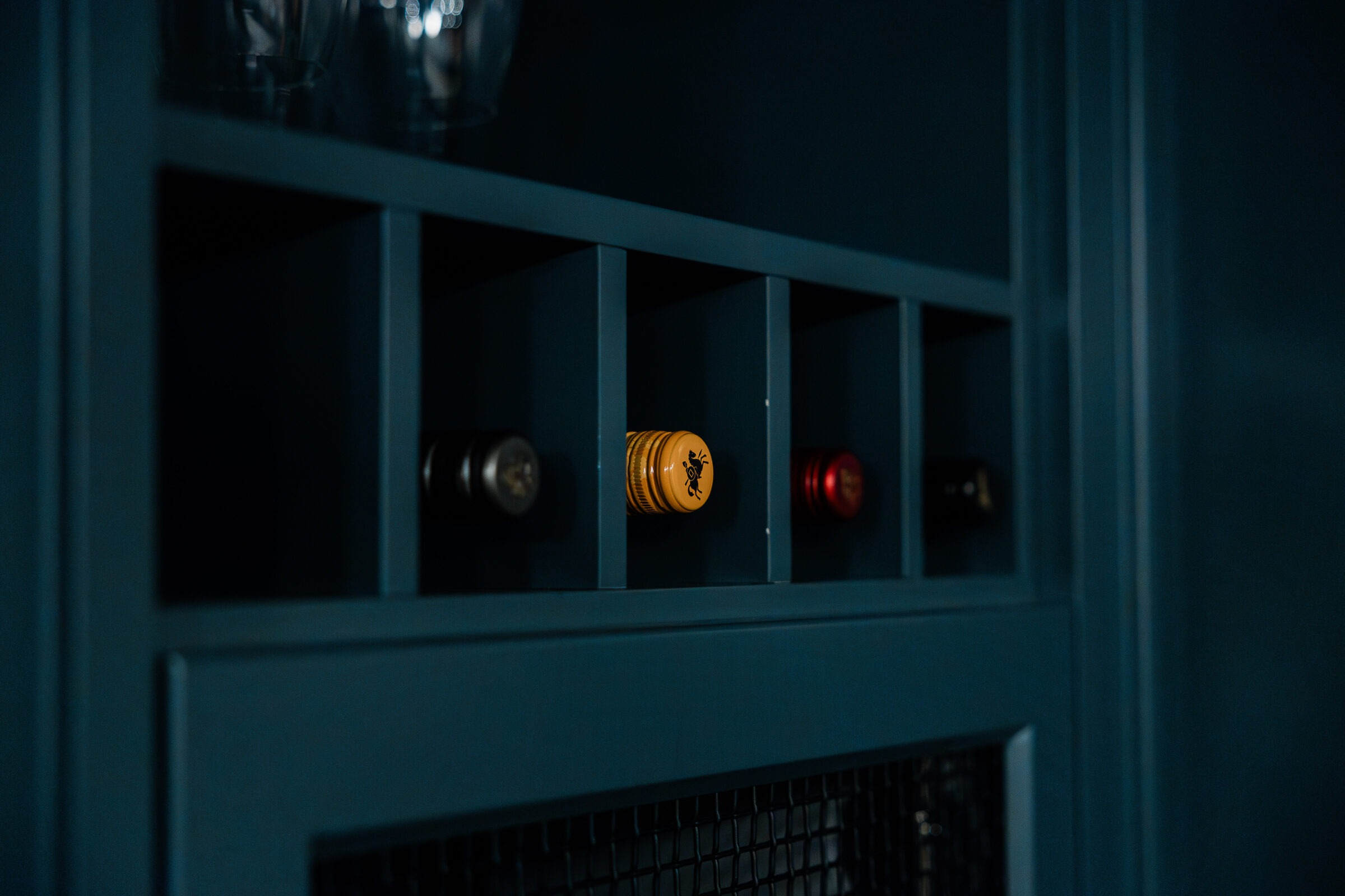 A dark, moody shot of a wine rack with bottles, characterized by a prominent golden cap centered in a blue cabinet, accompanied by subtle lighting.