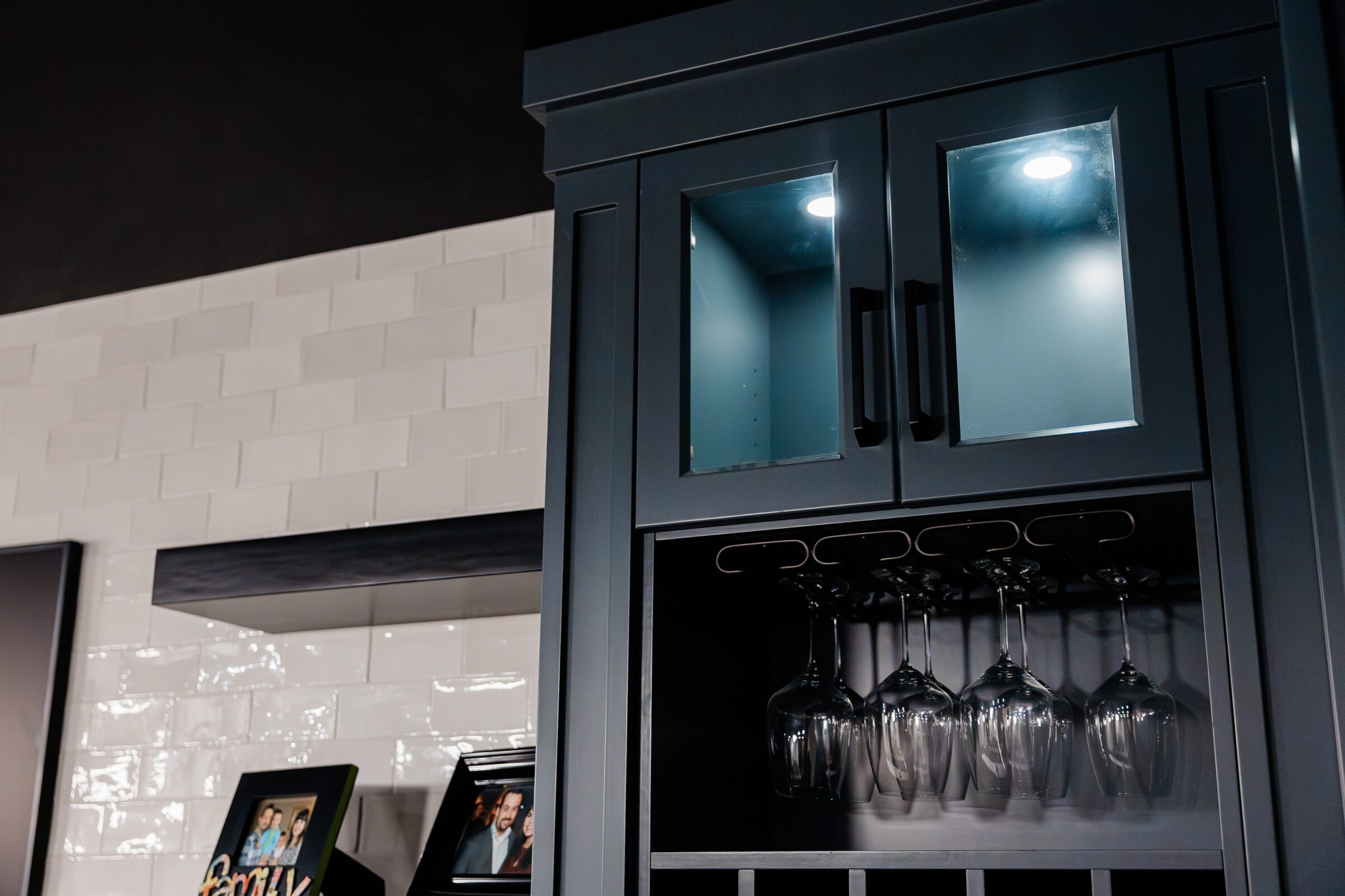 A modern kitchen cabinet with glass doors, translucent glassware hanging underneath, and a white subway tile backsplash. Framed photos are partially visible below.