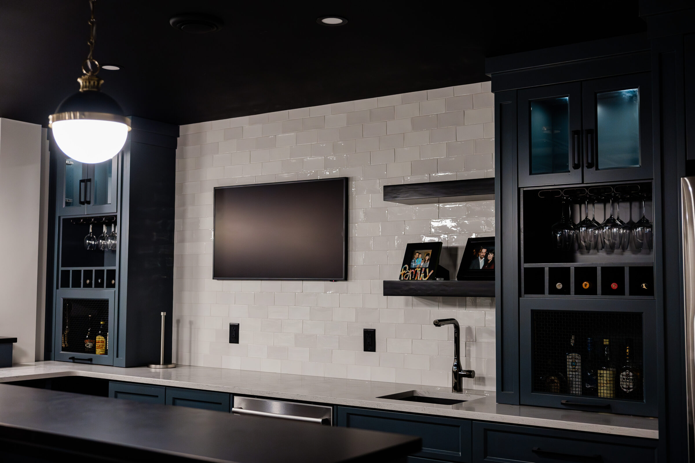 Modern kitchen with dark cabinets, white subway tile backsplash, stainless steel appliances, hanging glasses, and a ceiling pendant light above an island.