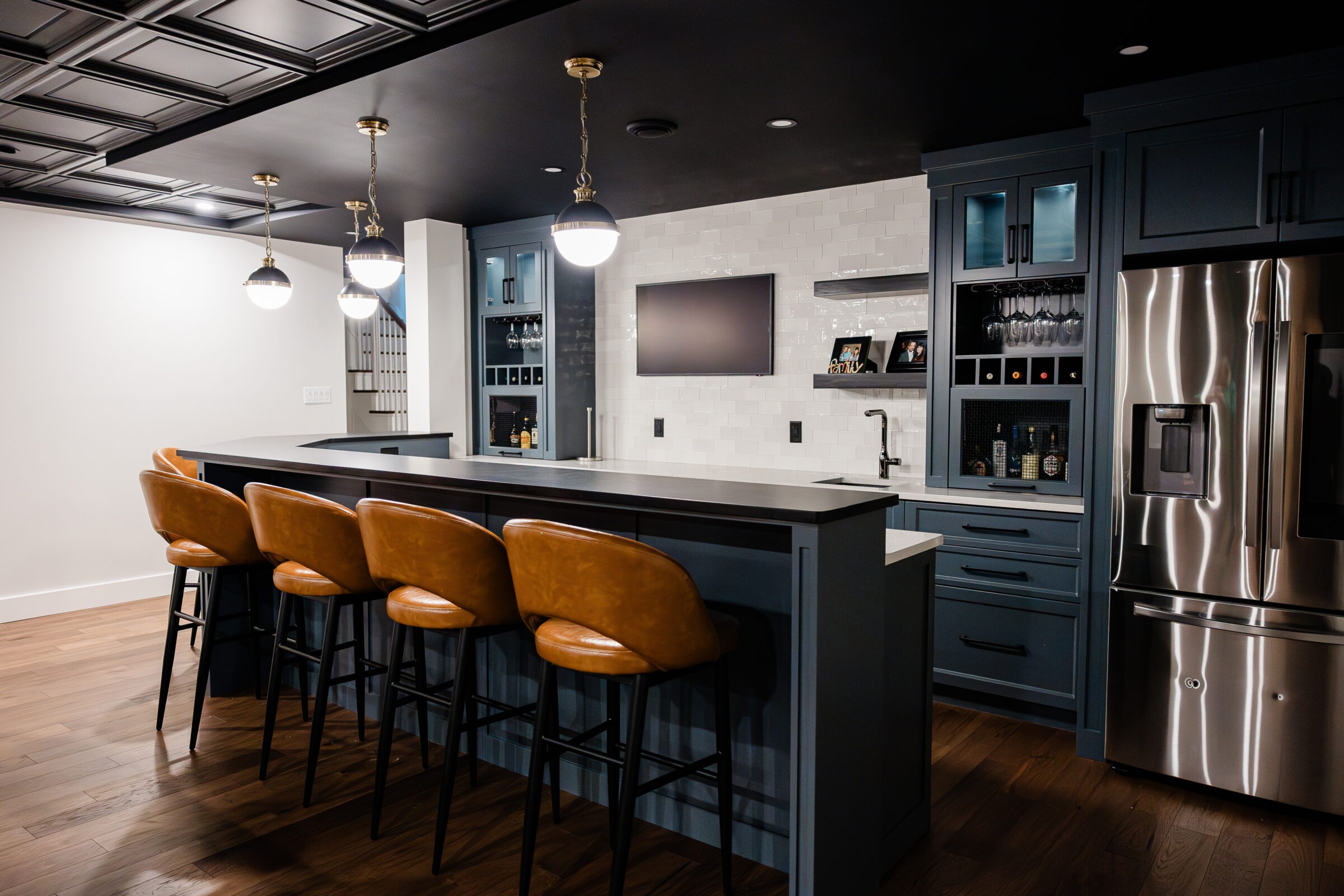 A modern kitchen with dark blue cabinetry, stainless steel fridge, hardwood floors, tan bar stools, white subway tiles, and gold-toned pendant lights.
