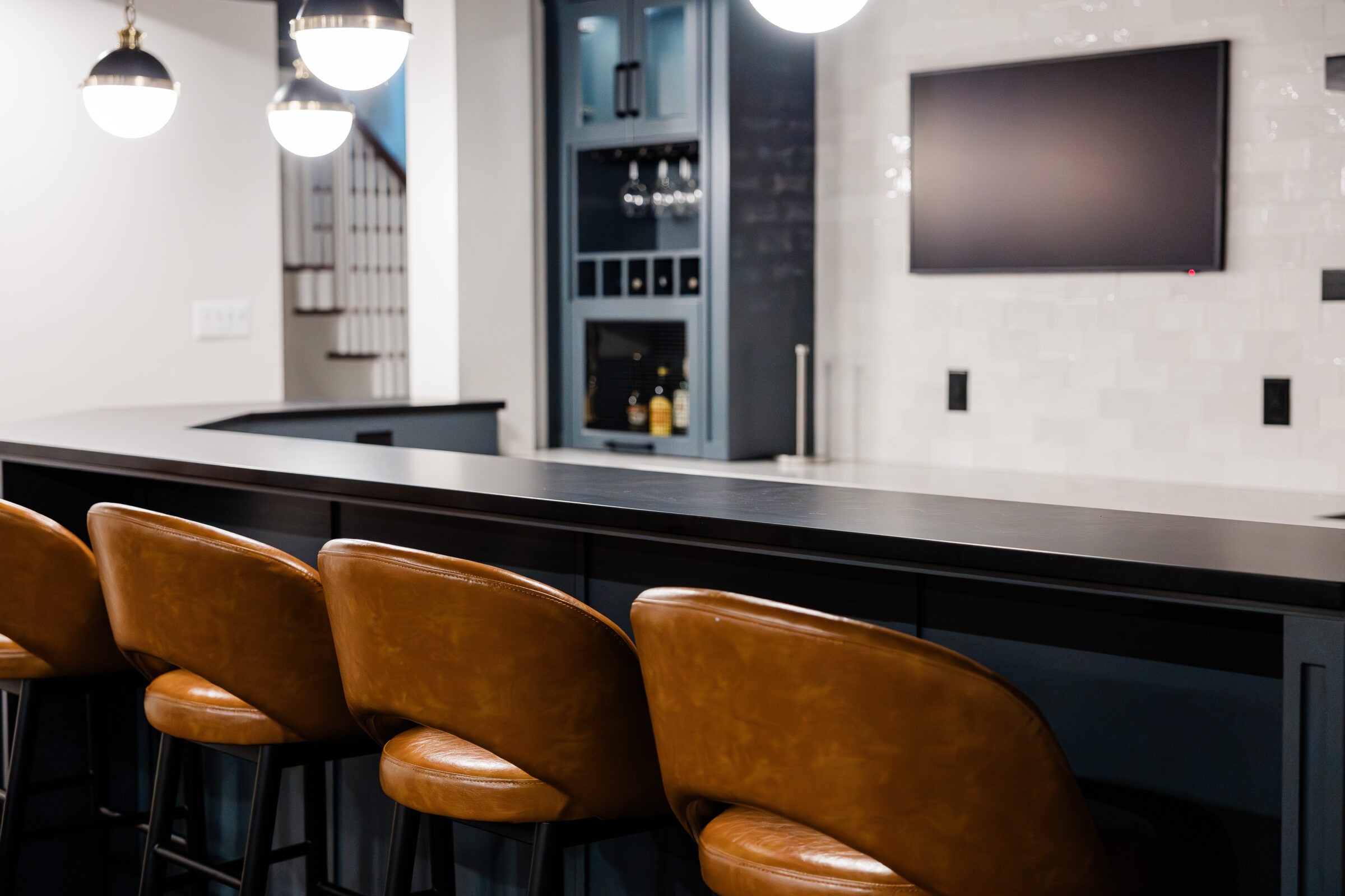 Modern kitchen interior with a sleek black countertop, tan leather bar stools, pendant lights, and a television on a subway-tiled wall.