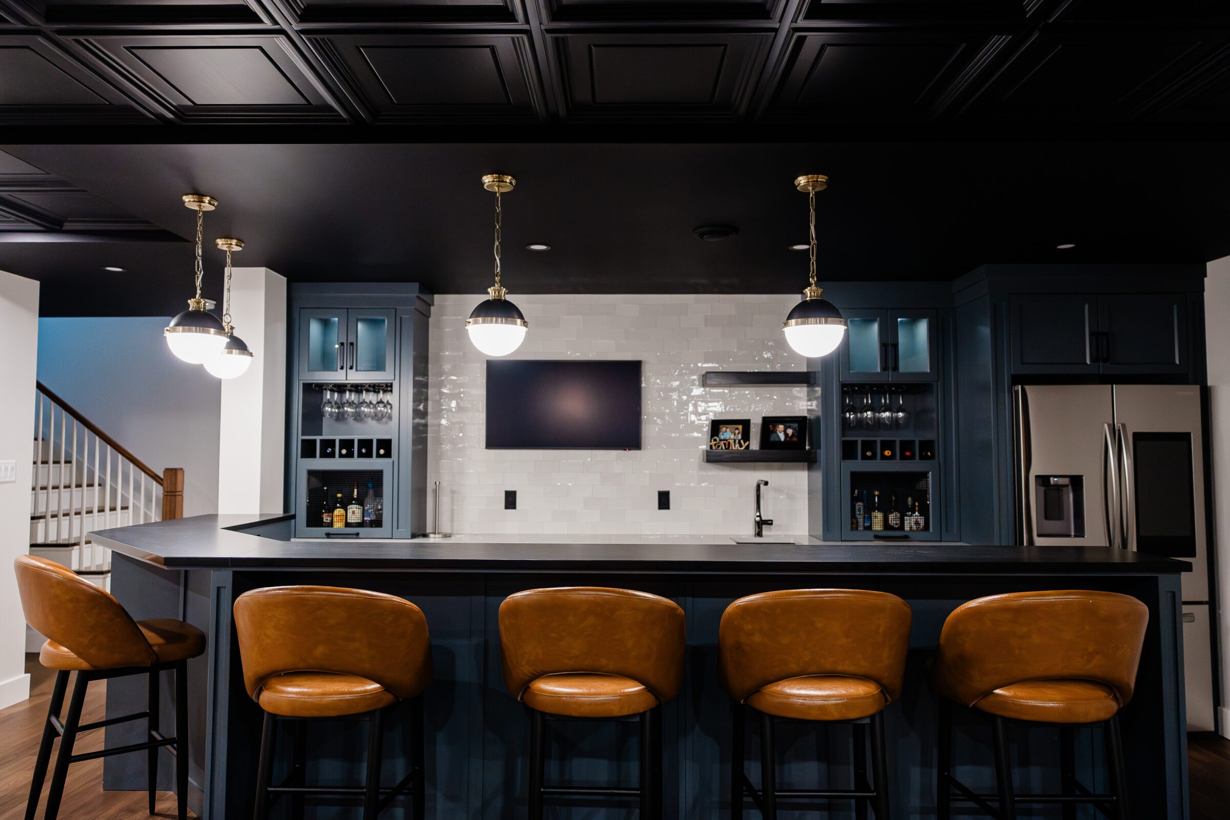 A stylish kitchen bar area with navy blue cabinetry, tan leather barstools, pendant lights, white backsplash, and modern appliances under a dark ceiling.