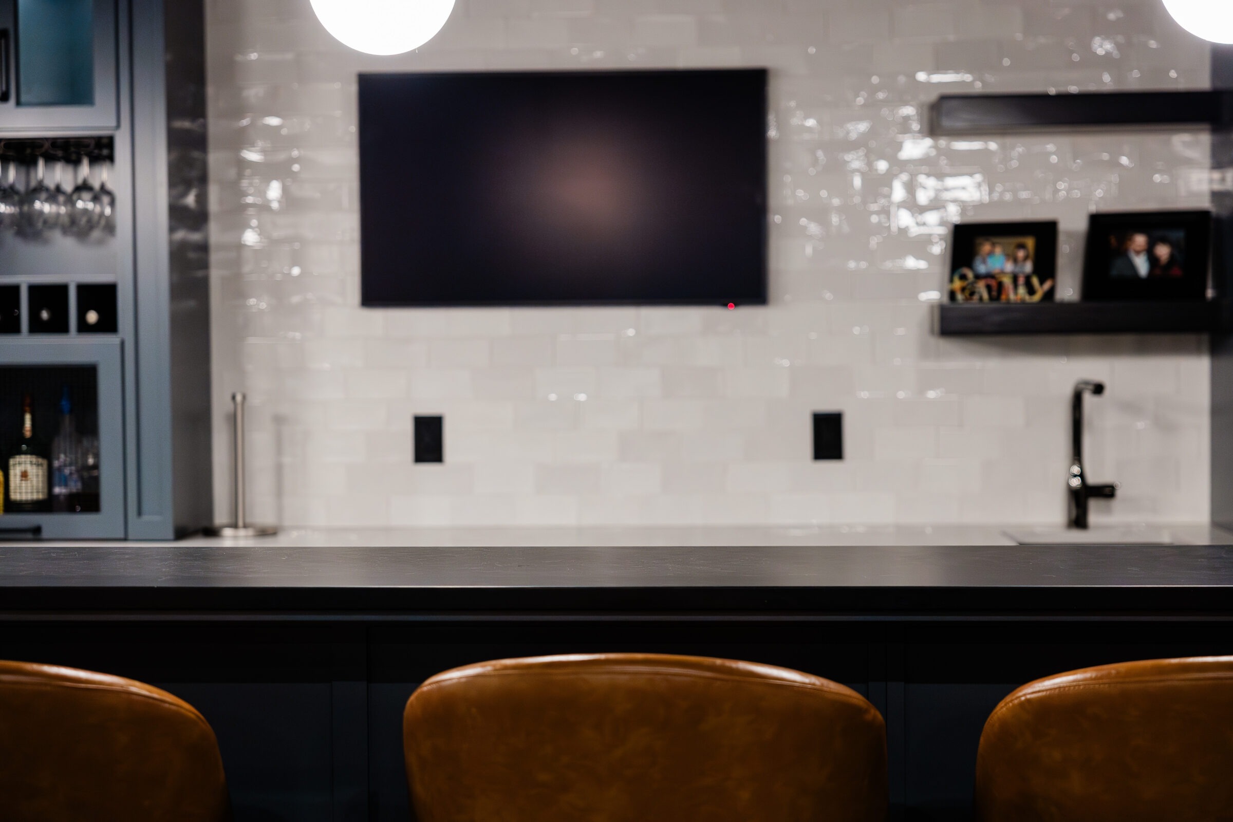 An elegant kitchen with a dark counter, three leather stools, a tiled backsplash, and a mounted TV. Cabinets and a shelf hold glassware and bottles.