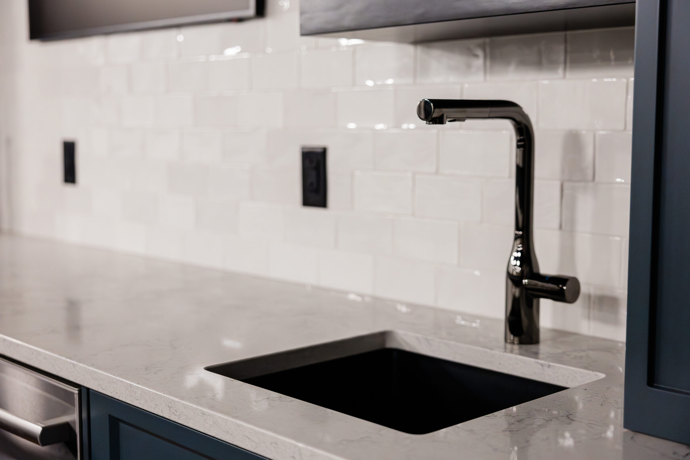 A modern kitchen with white subway tiles, a gray countertop, a black undermount sink, and a matte black faucet with corresponding black switch plates.