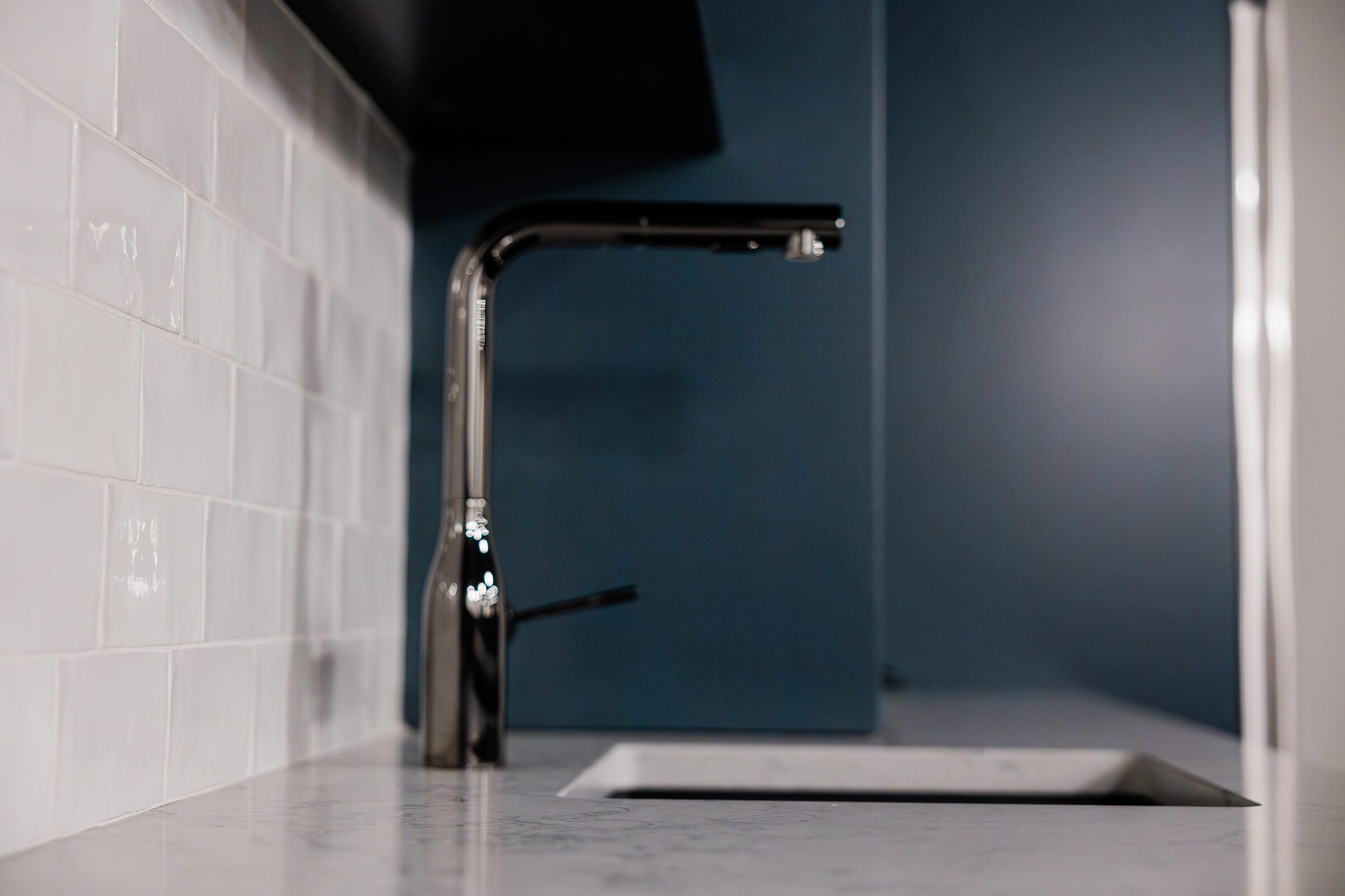 This image shows a close-up of a modern kitchen sink with a stainless steel faucet against a backdrop of white tile and dark blue walls.