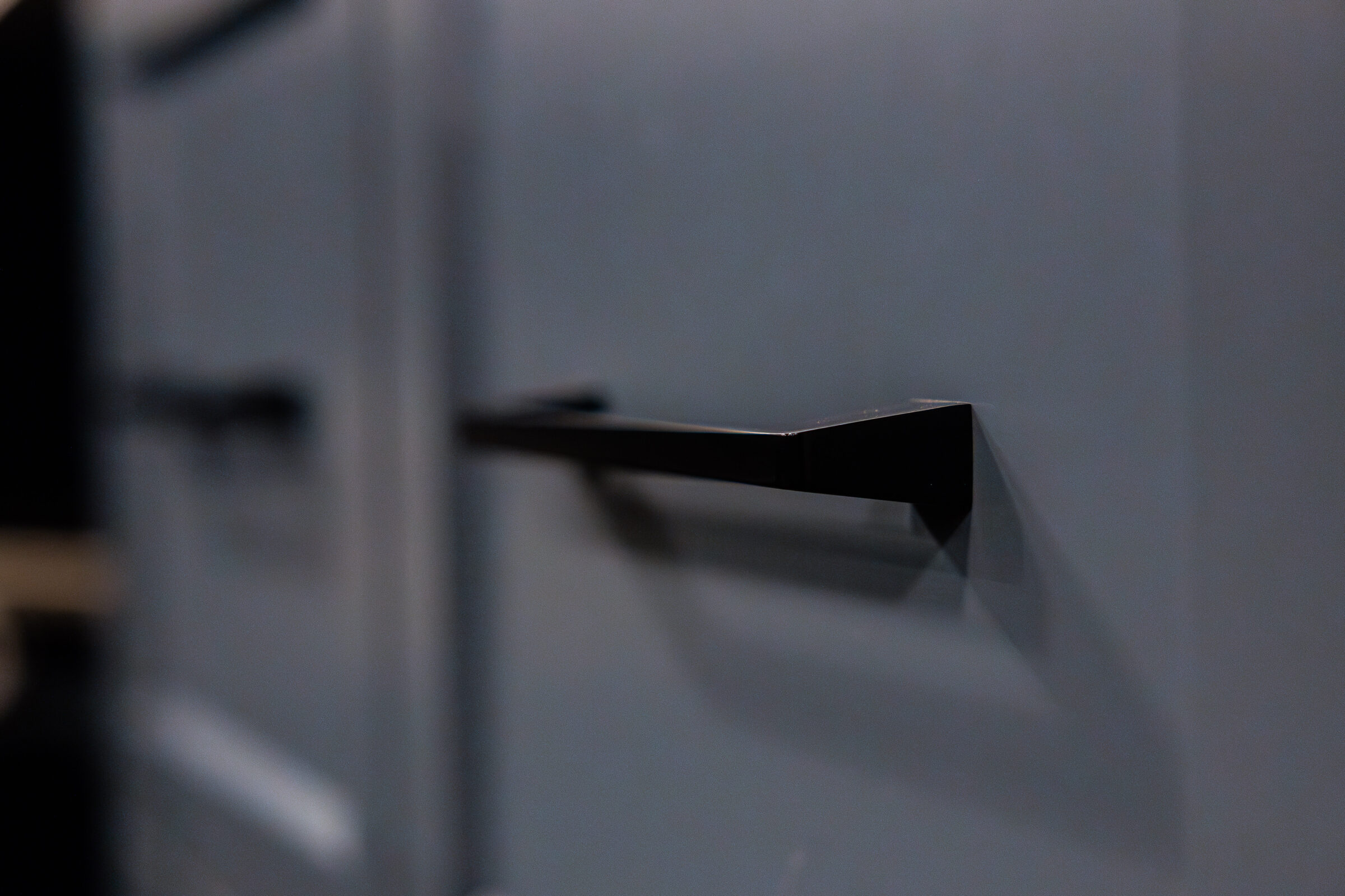 The image shows a close-up of a modern black cabinet handle against a blurred blue background, emphasizing the simplicity and elegance of the design.
