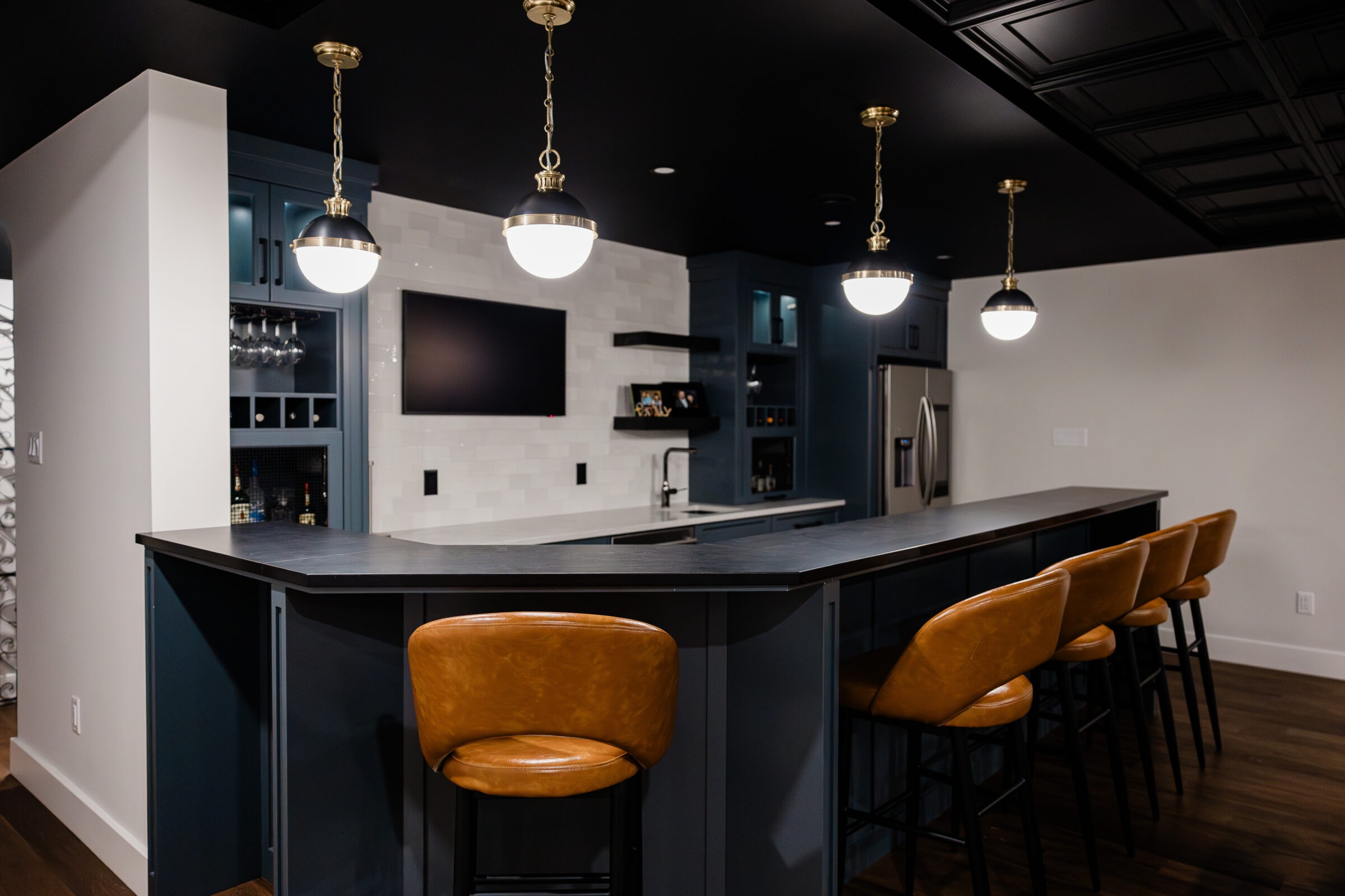 A modern kitchen with dark cabinetry, a large island with brown leather stools, pendant lights, and stainless steel appliances, featuring a wine rack.