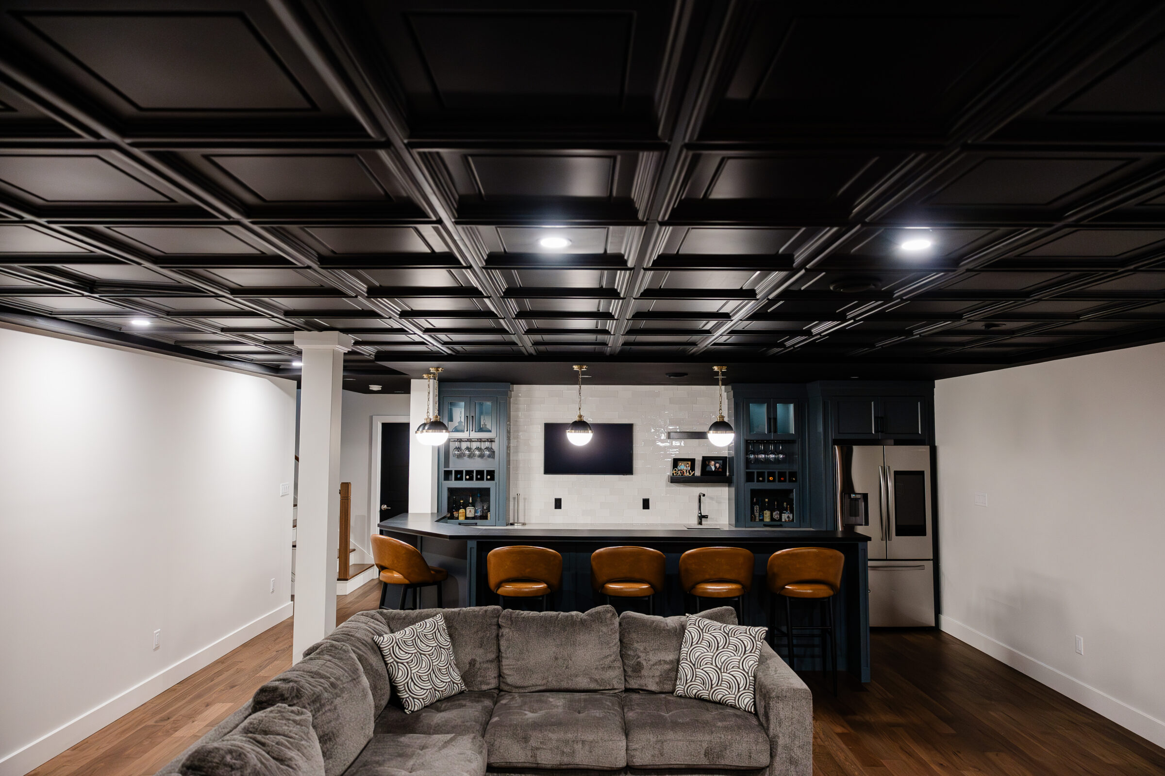 A modern kitchen with dark cabinetry and a tile backsplash, featuring bar stools, pendant lighting, and a coffered ceiling with recessed lights.