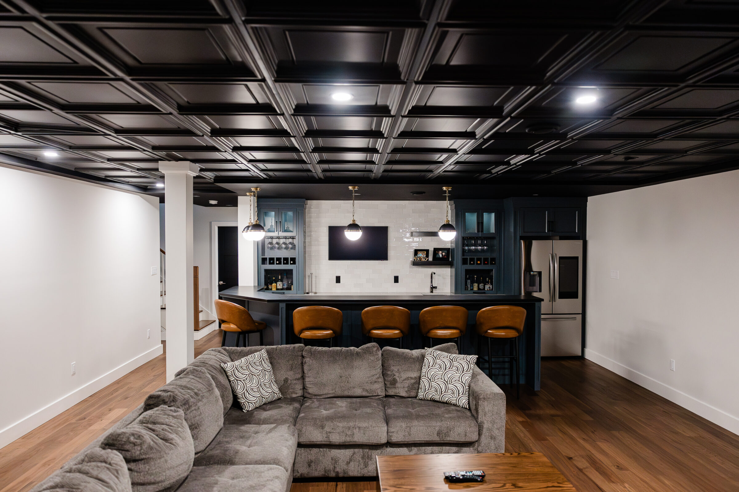An elegant interior space featuring a black coffered ceiling, hardwood floors, a gray sectional sofa, and a kitchen with bar stools.