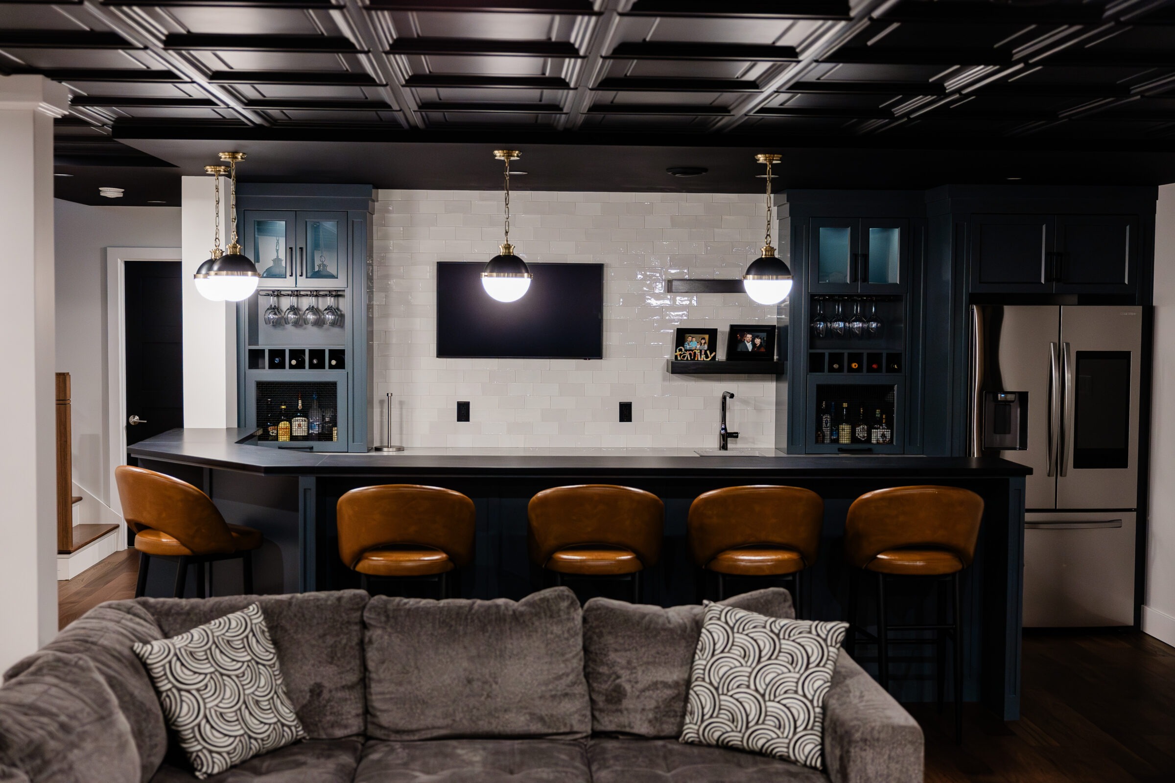 A stylish kitchen bar with a black ceiling, modern pendant lighting, dark cabinets, and brown leather stools, complemented by a cozy gray sectional sofa.