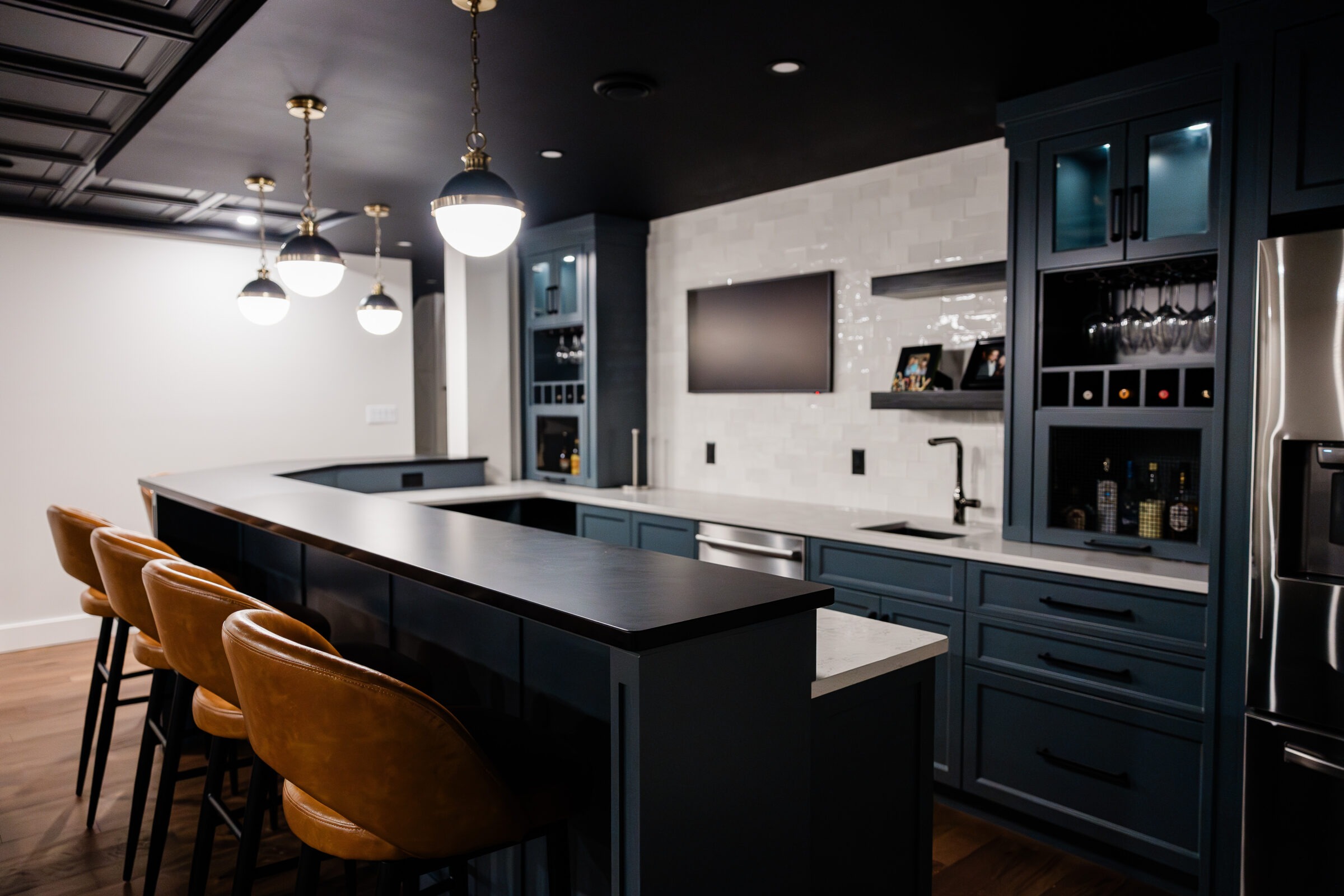Modern kitchen interior showcasing a dark countertop island with tan stools, blue cabinetry, stainless steel appliances, and pendant lighting.