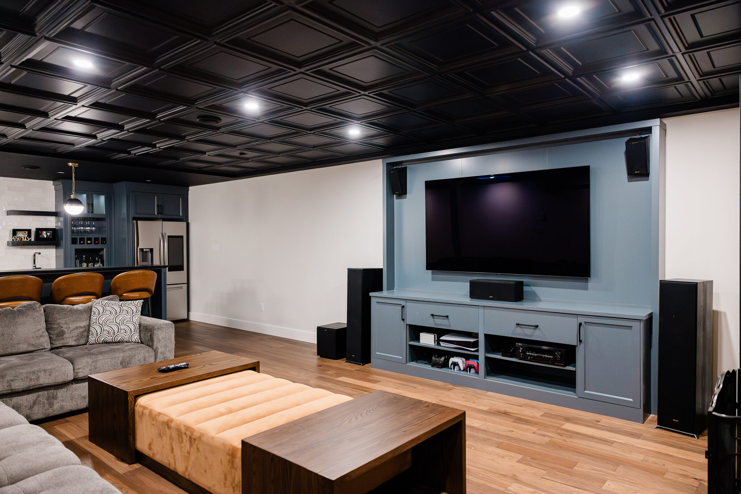 Modern living room with a sectional sofa, a wooden coffee table, herringbone floor, and a large TV on a blue media console under a dark ceiling.