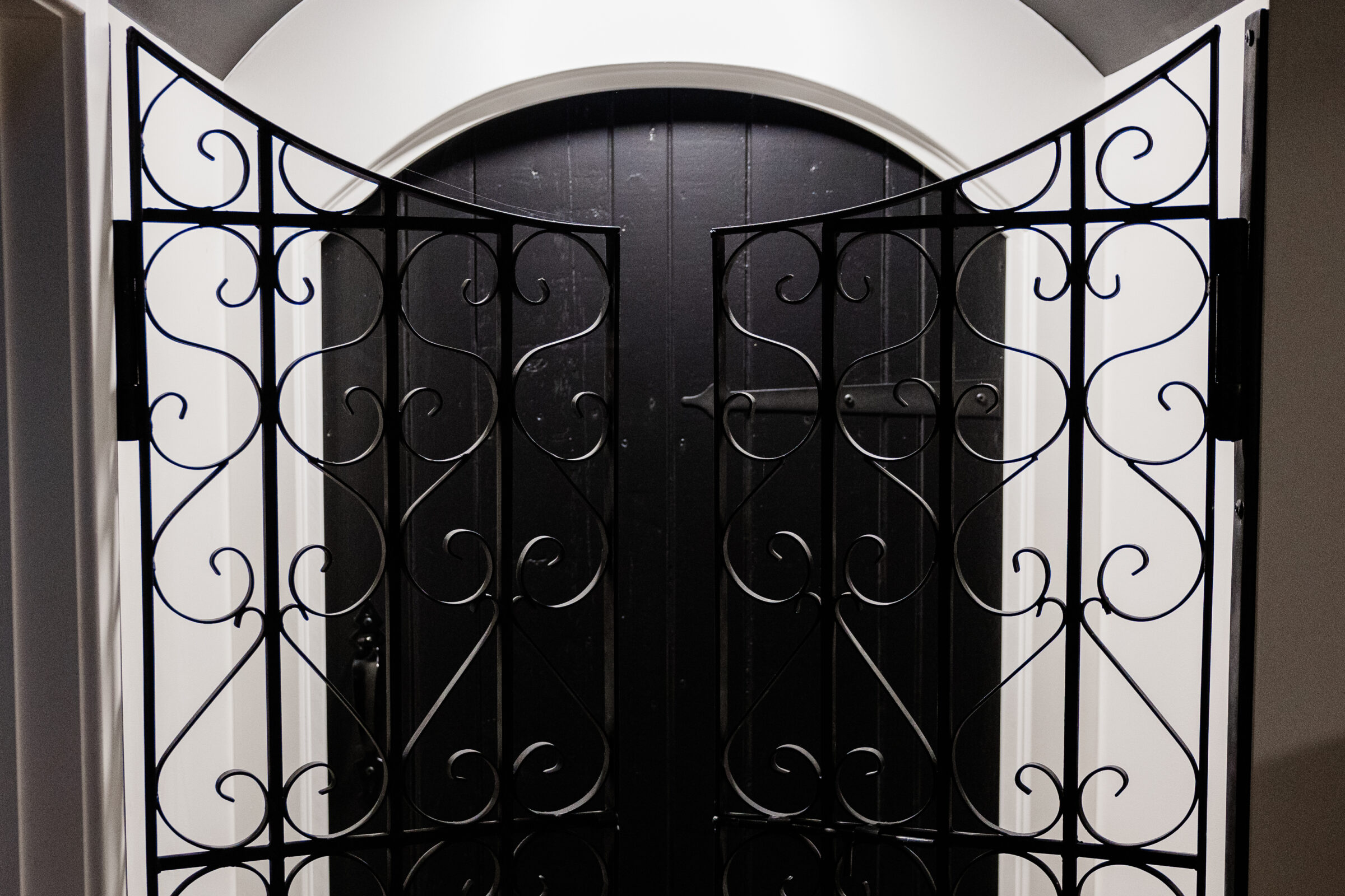 This image shows a black wrought iron gate with ornate scroll designs, closed in front of a dark arched doorway with white trim.