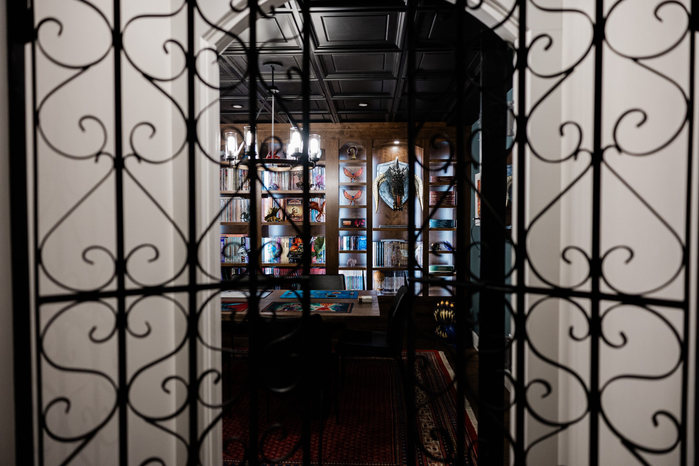 This image shows an ornate room viewed through decorative wrought iron gates, featuring a bookshelf, eclectic decorations, and a chandelier, creating an inviting atmosphere.