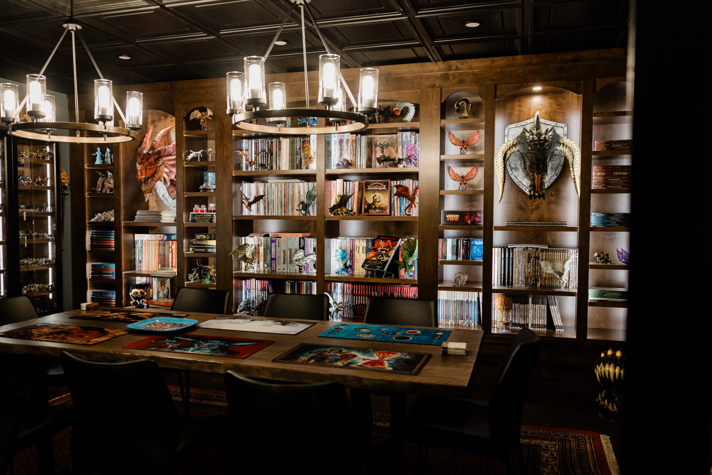 A cozy room with bookshelves filled with books and collectibles, a table with artwork, circular pendant lighting, and a dragon sculpture on the wall.