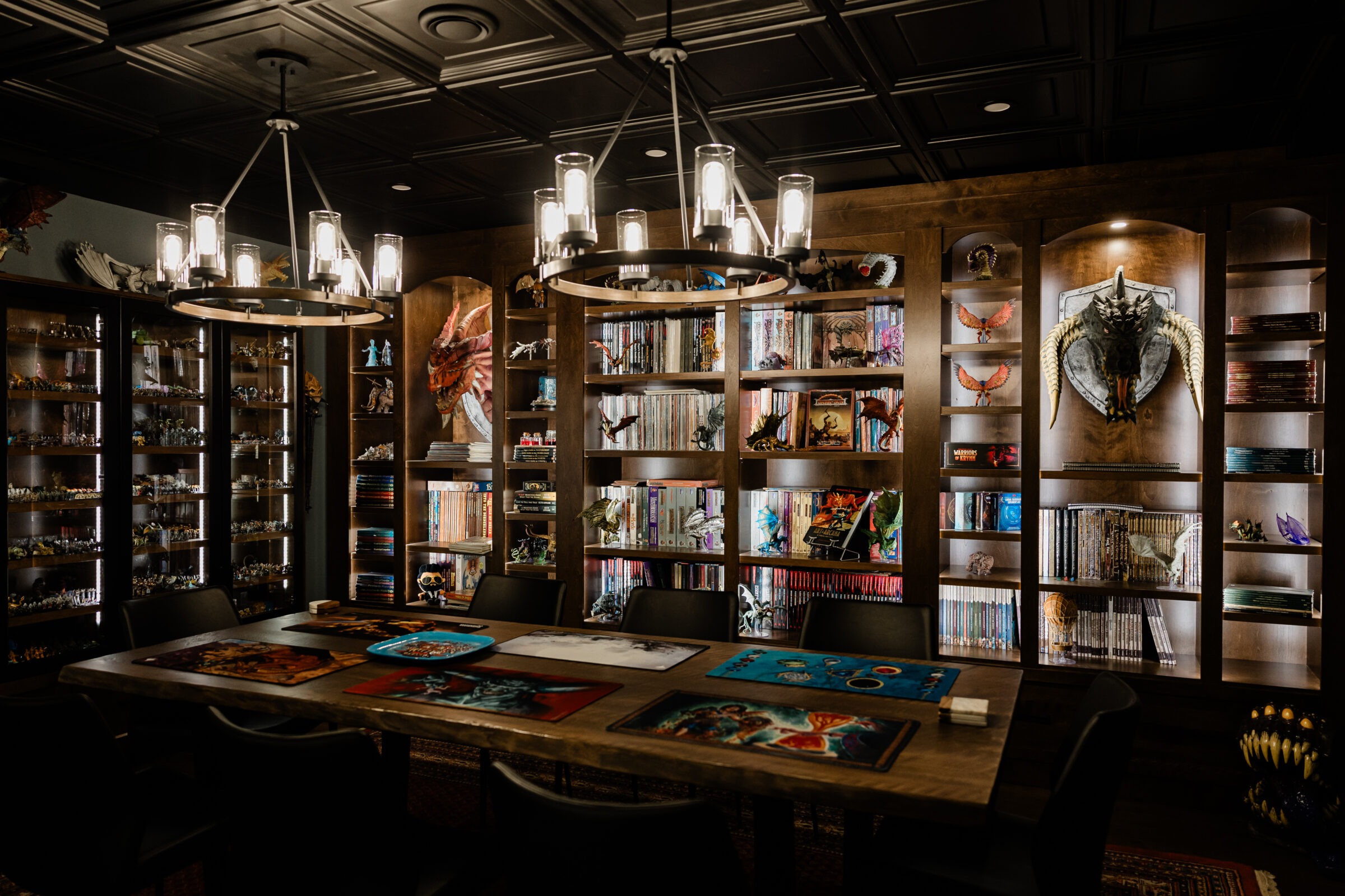 A dimly lit room adorned with shelves of books, figures, and artwork. Central table displays colorful board game materials, creating an inviting gaming space.