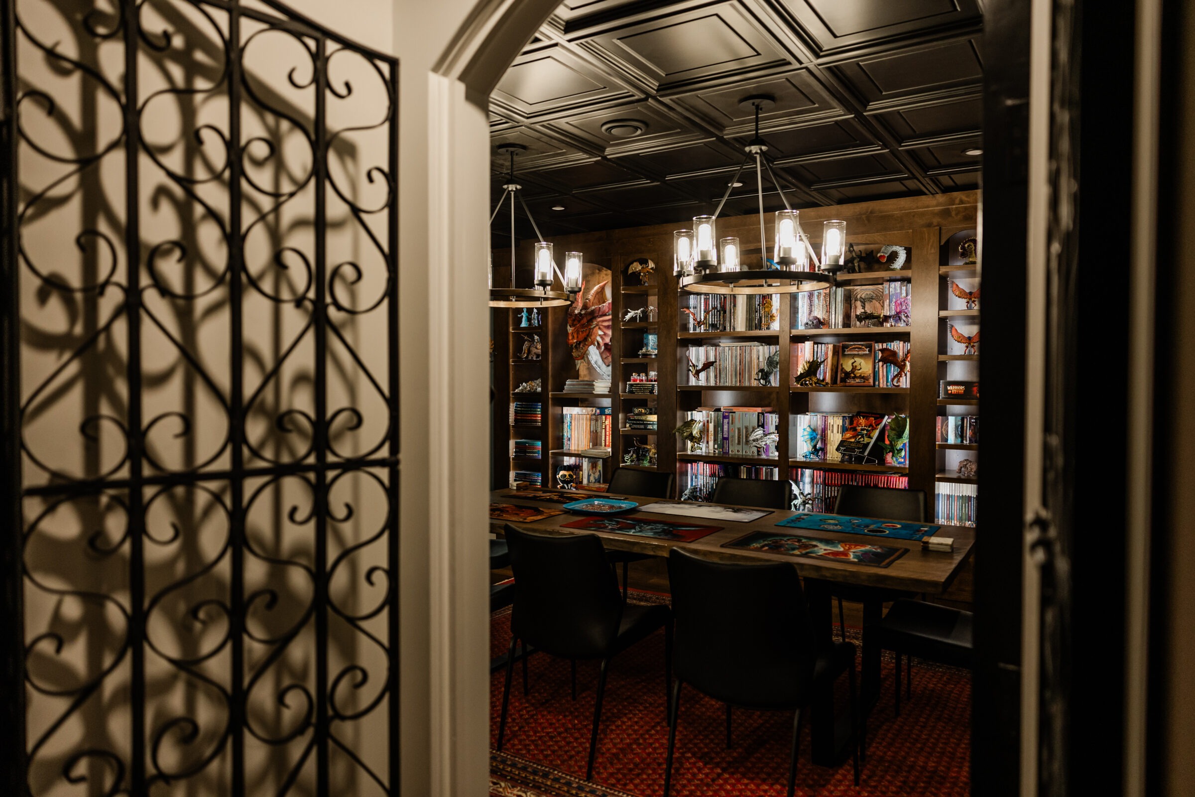 An elegant room with bookshelves, hanging lights, and a table with chairs. A decorative metal gate opens to the warmly lit, cozy interior space.