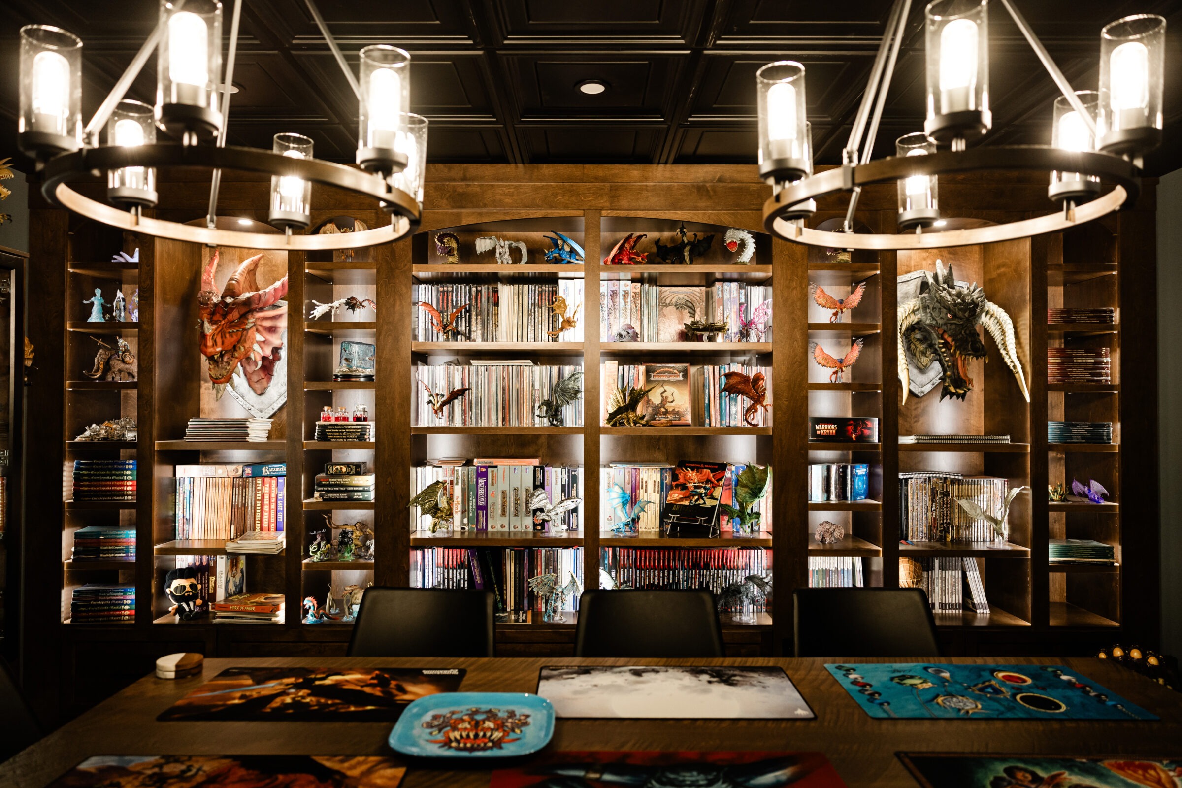 A cozy room with bookshelves filled with books, figurines, and fantasy artwork. A large table with gaming mats sits under modern chandeliers.