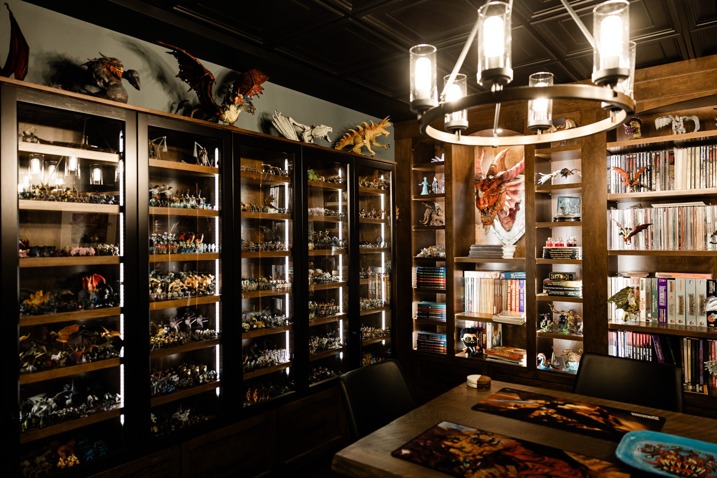 A dimly lit room with wooden bookshelves filled with colorful miniatures and books, a chandelier above, and fantasy-themed decor adorning the walls.