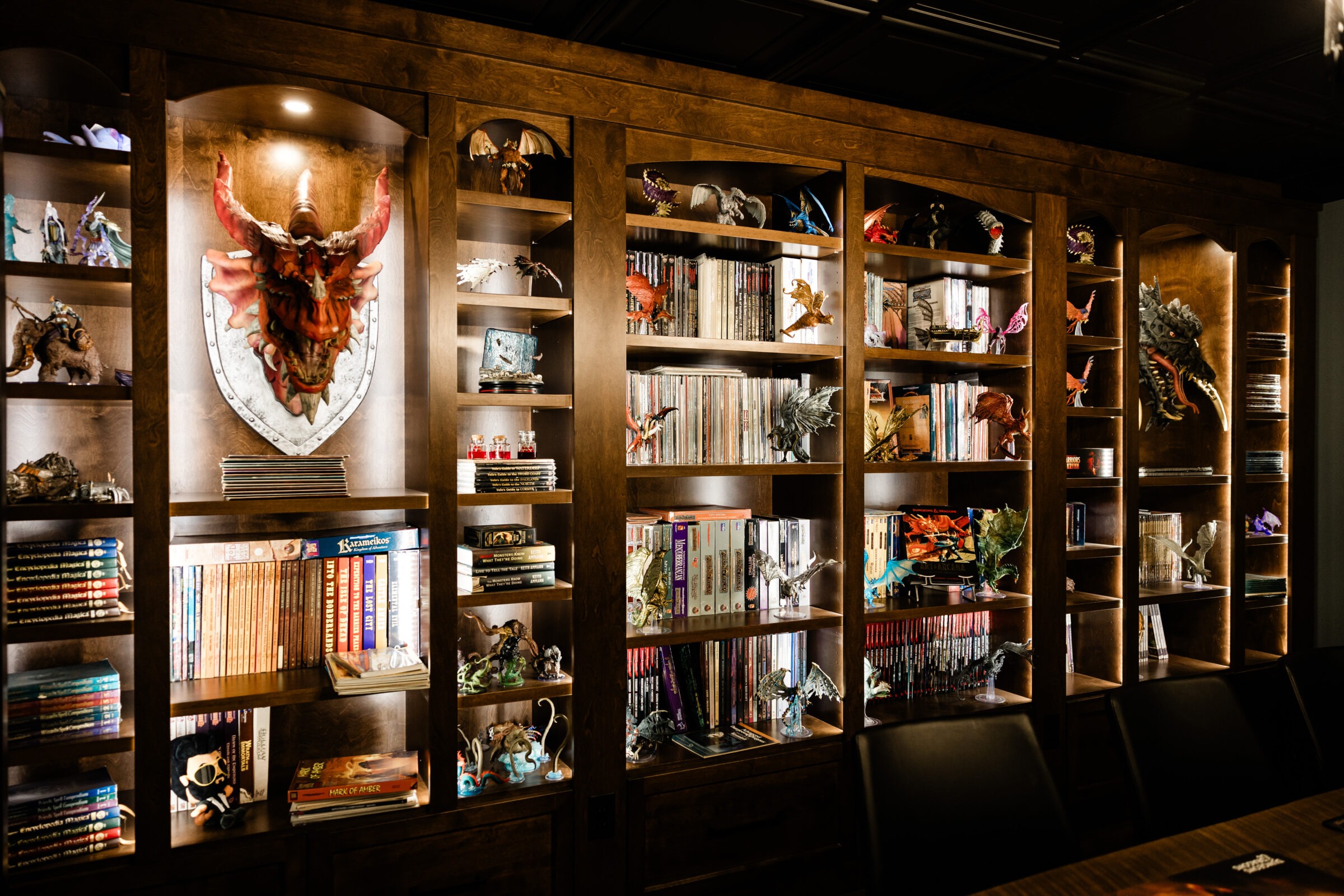 A large wooden bookshelf is filled with books and various colorful fantasy and dragon figurines. A shield with a dragon skull serves as a centerpiece.