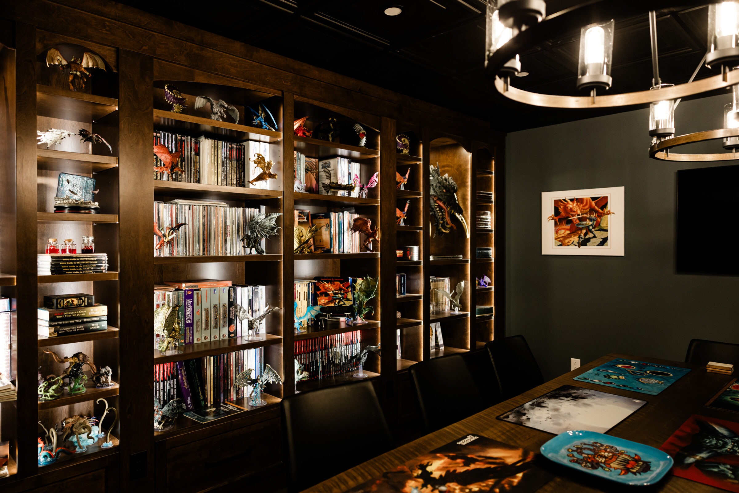 A well-lit room with a large wooden bookshelf filled with books, colorful figurines, and collectibles. A dark table with artistic mats is centered below a chandelier.