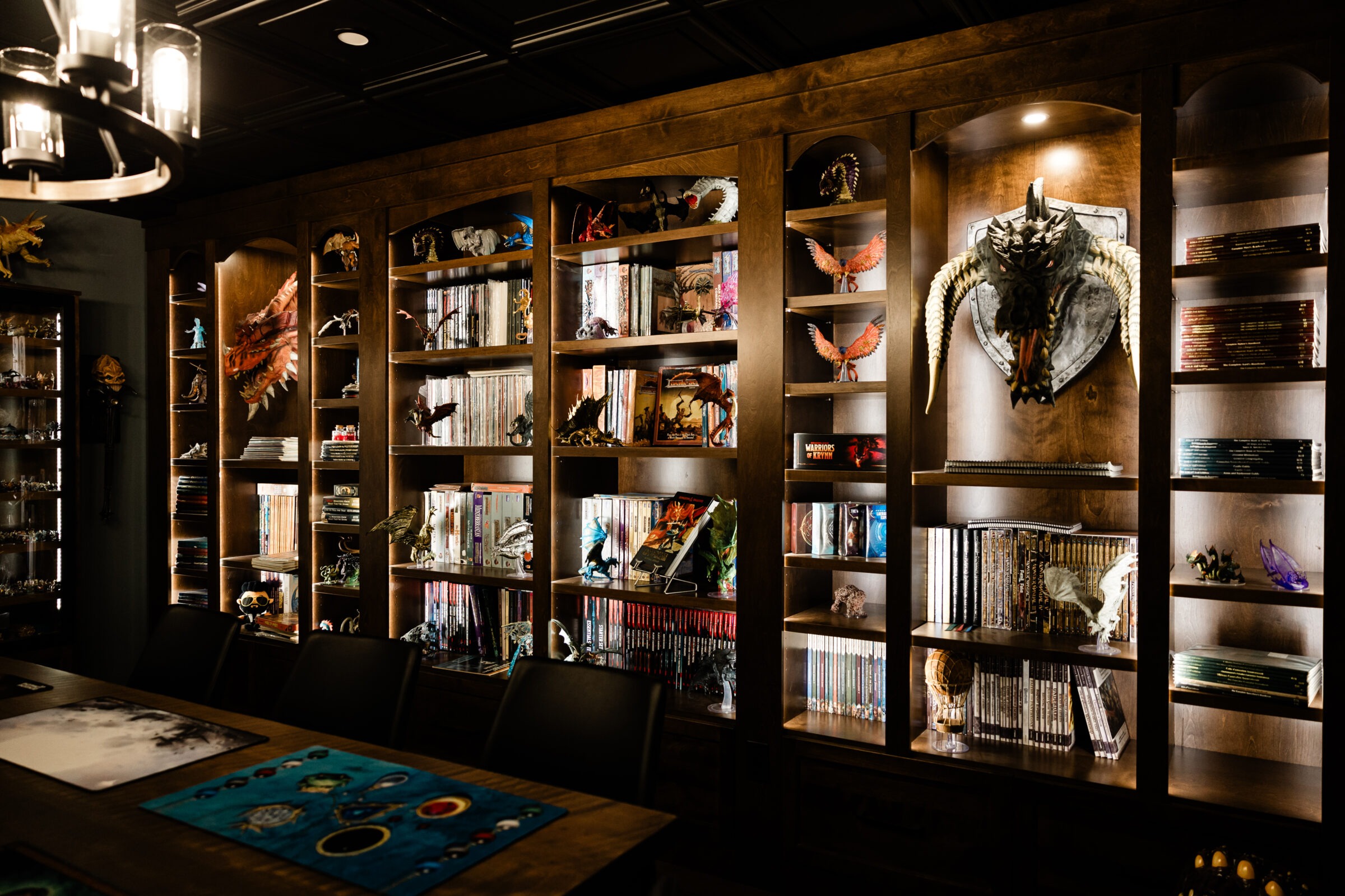 An interior with wooden shelves filled with books, figurines, and collectibles. A dragon head mount is central, above a table with a colorful mat.