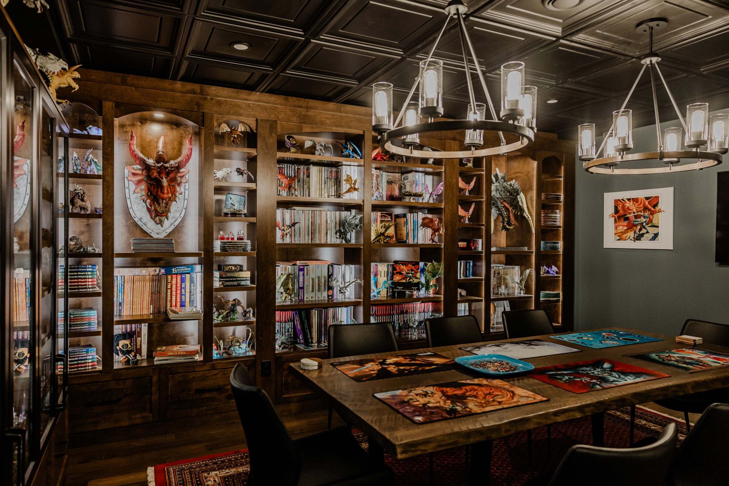 A cozy room with wooden bookshelves filled with books and figurines, tables adorned with colorful artwork, and modern hanging light fixtures above.