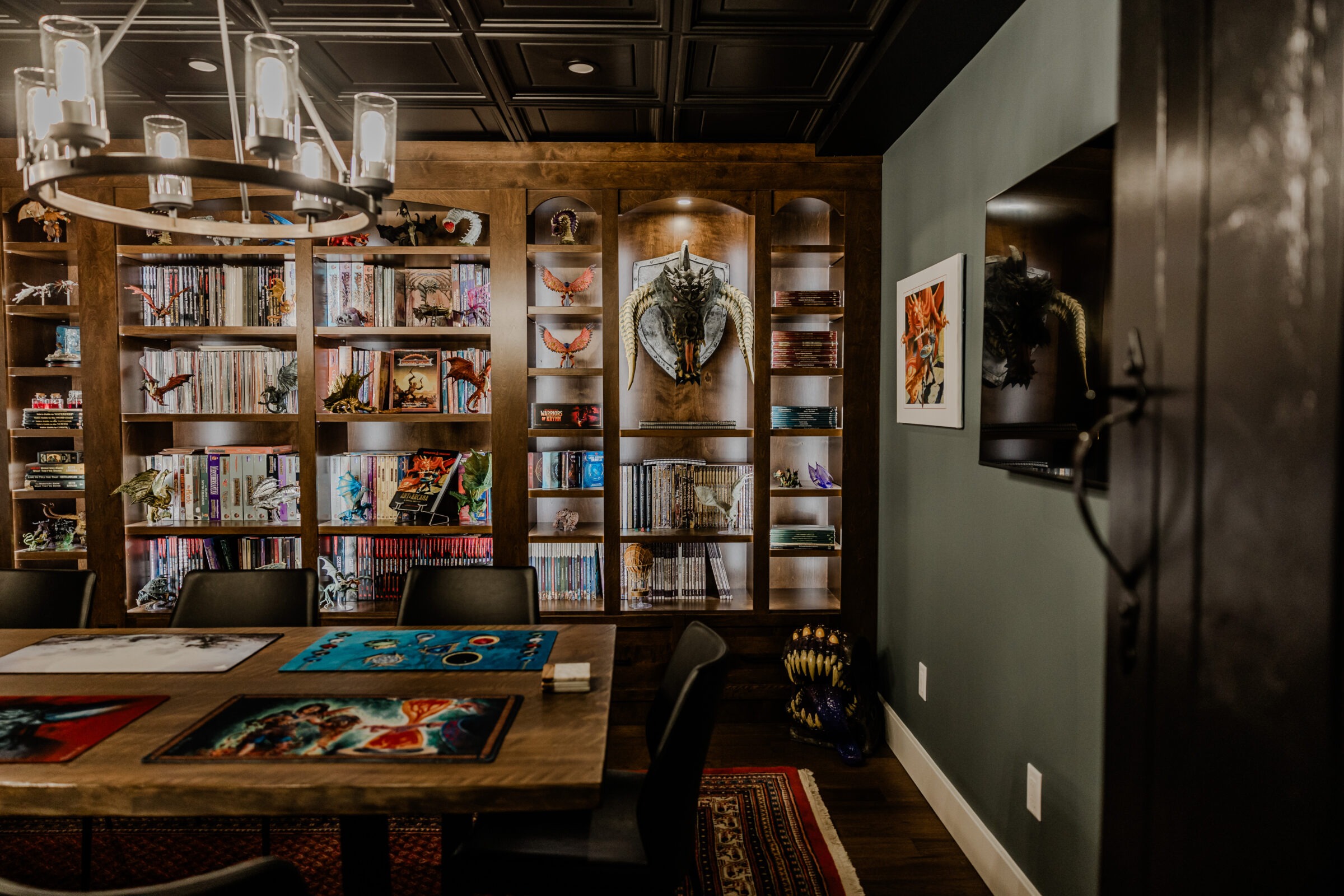 A dimly lit room with wooden bookshelves filled with books and figurines, a table with artwork, a chandelier above, and dragon-themed wall decor.