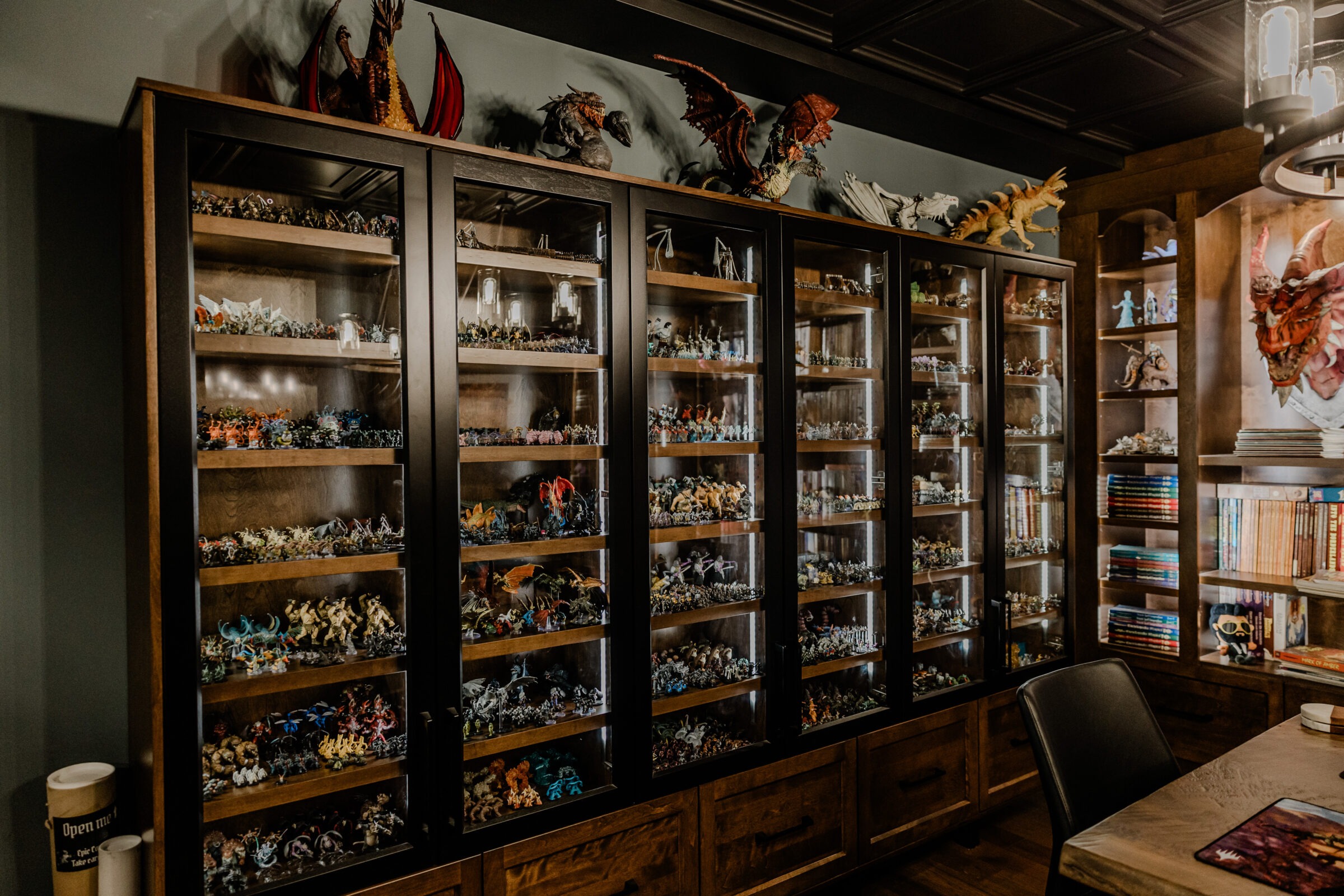 A dim wood-paneled room with large glass display cases, filled with an extensive collection of miniature fantasy figures and dragons on top.