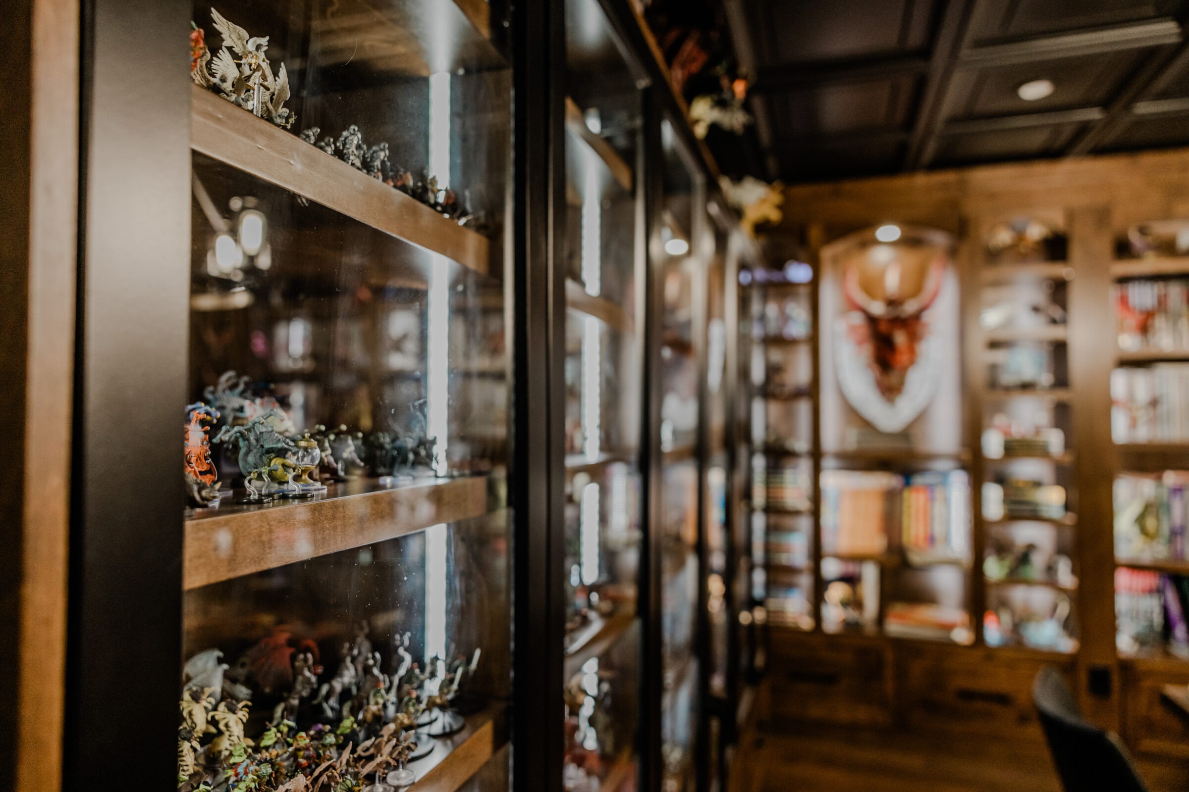The image showcases a cozy interior with a glass cabinet displaying a collection of miniatures. A bookshelf and decor, including a wall-mounted stag head, are visible.