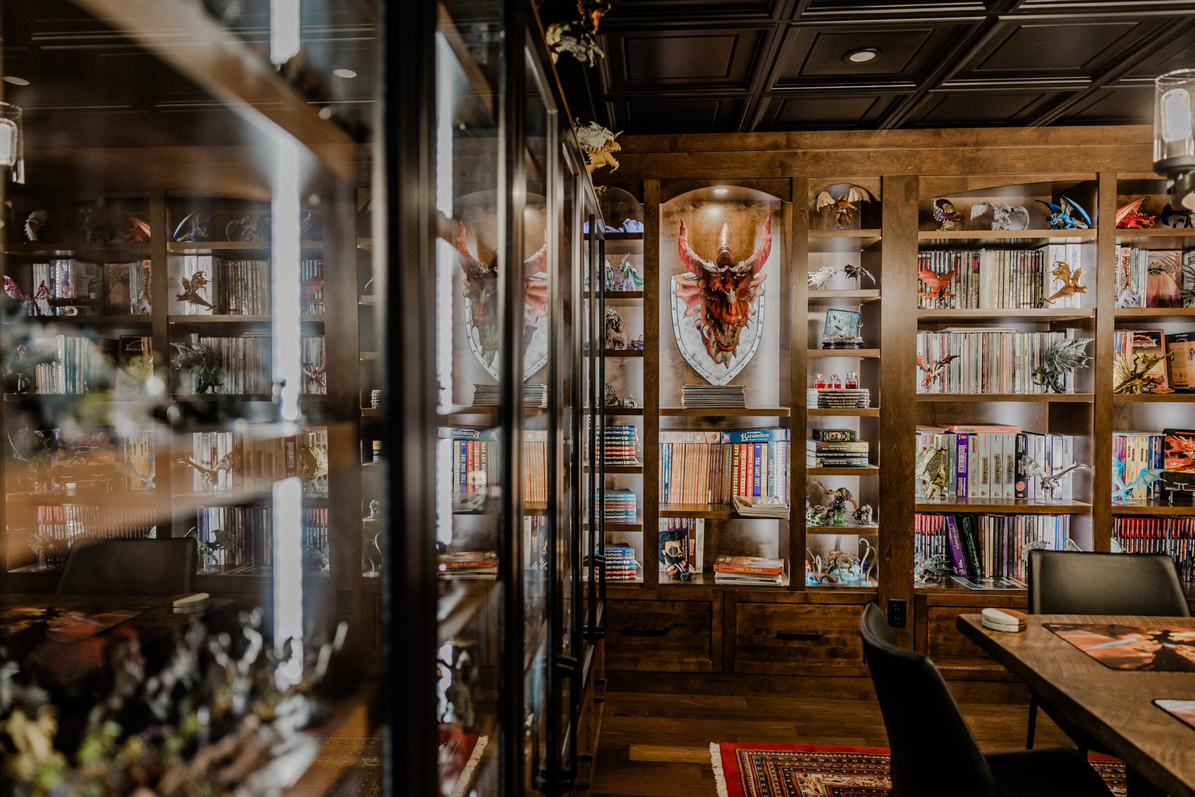 The image shows a cozy, book-filled room with wooden floors and shelves, various ornaments, a large decorative shield, and a glimpse of a person.
