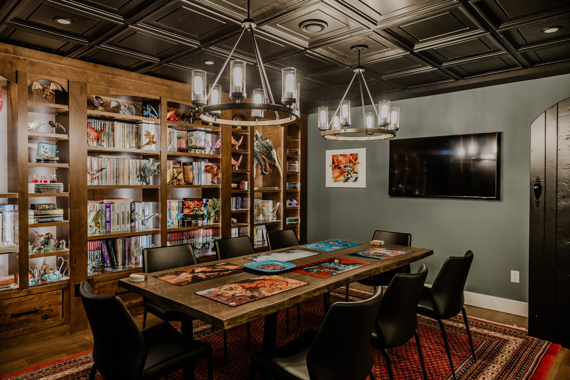 A cozy room featuring bookshelves filled with books, board games laid out on a wooden table, modern hanging lights, a large TV, and a dark ceiling.