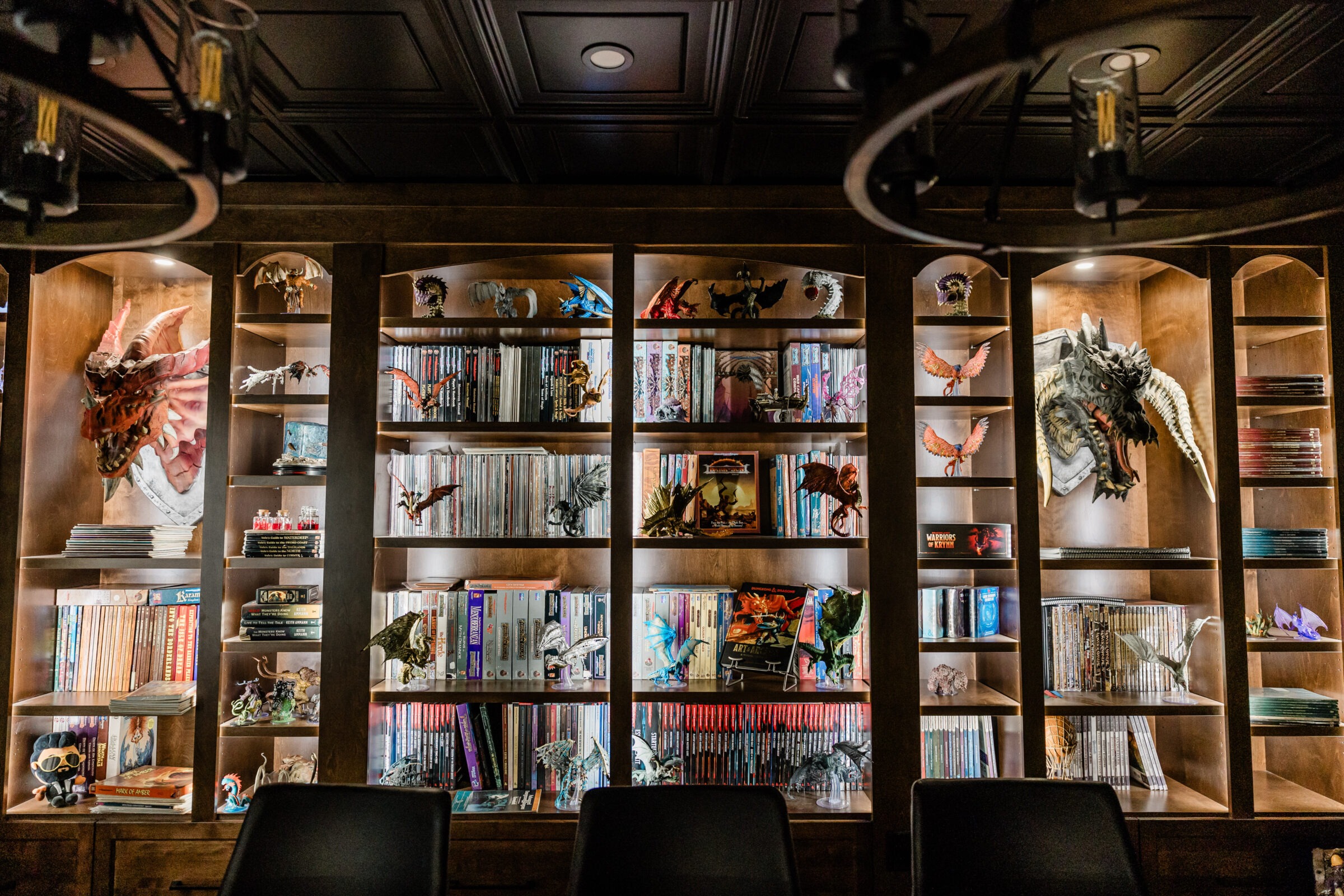 A room with wooden shelving displaying numerous books, collectible figures, and dragon models under warm lighting with visible hanging light fixtures.