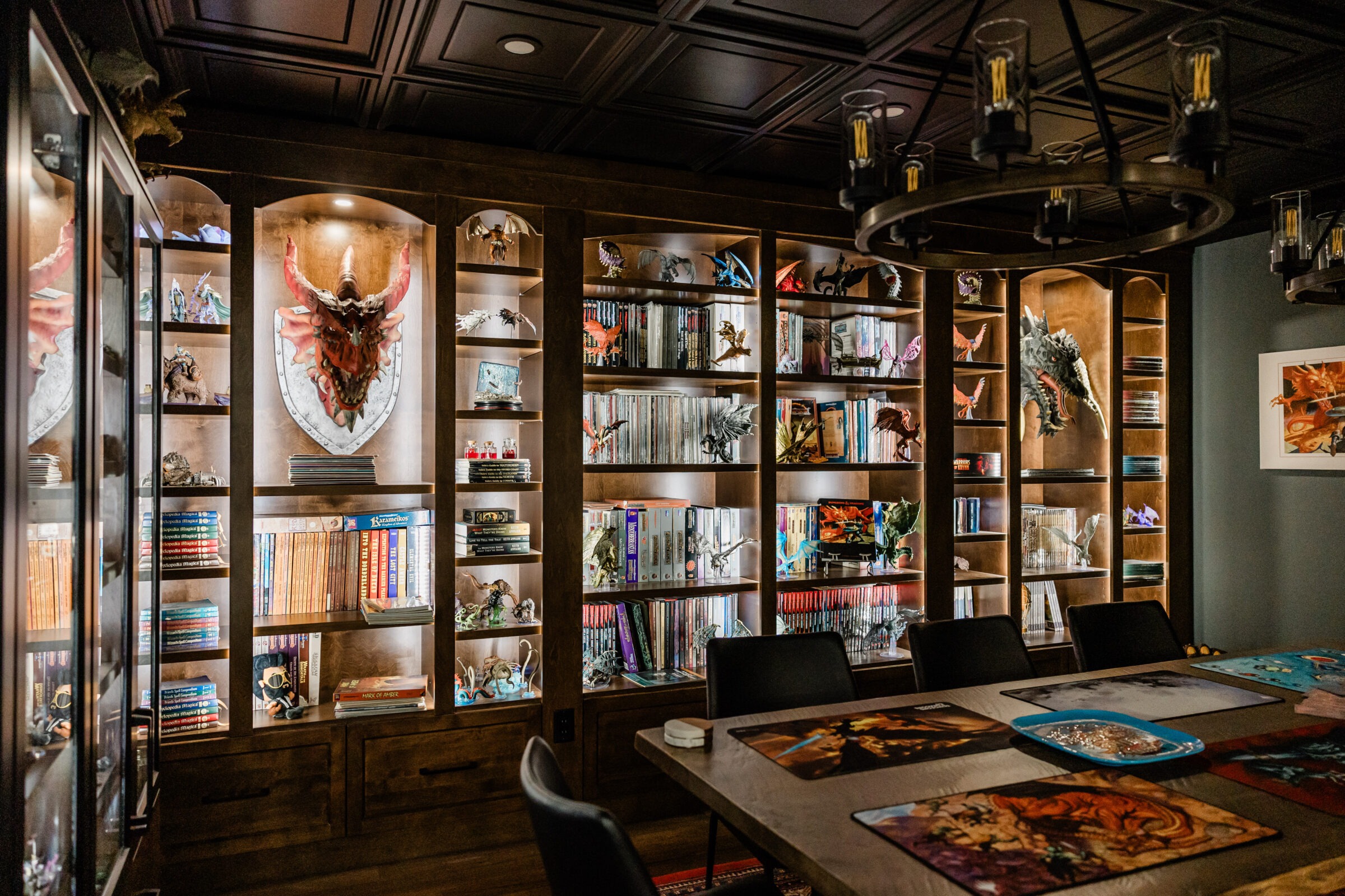 A well-lit room with wood-paneled walls features bookshelves full of books, collectibles, and framed artwork, above a table set with gaming mats.
