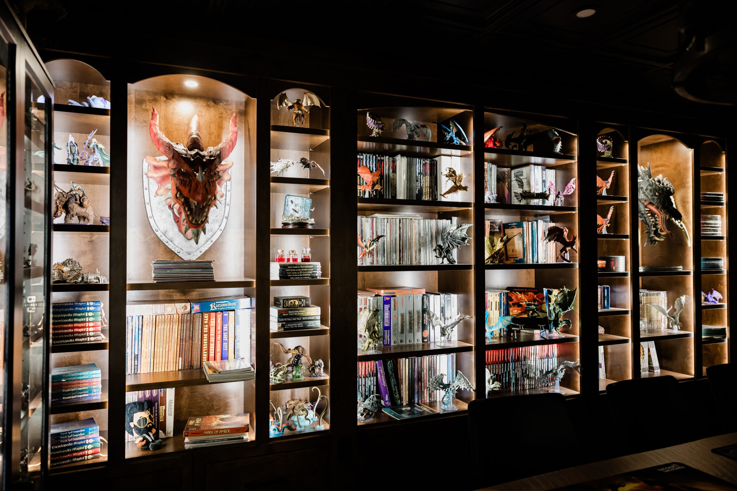 A well-lit bookshelf displays an assortment of books, intricate dragon figures, and fantasy-themed collectibles in a cozy, dark-toned room.