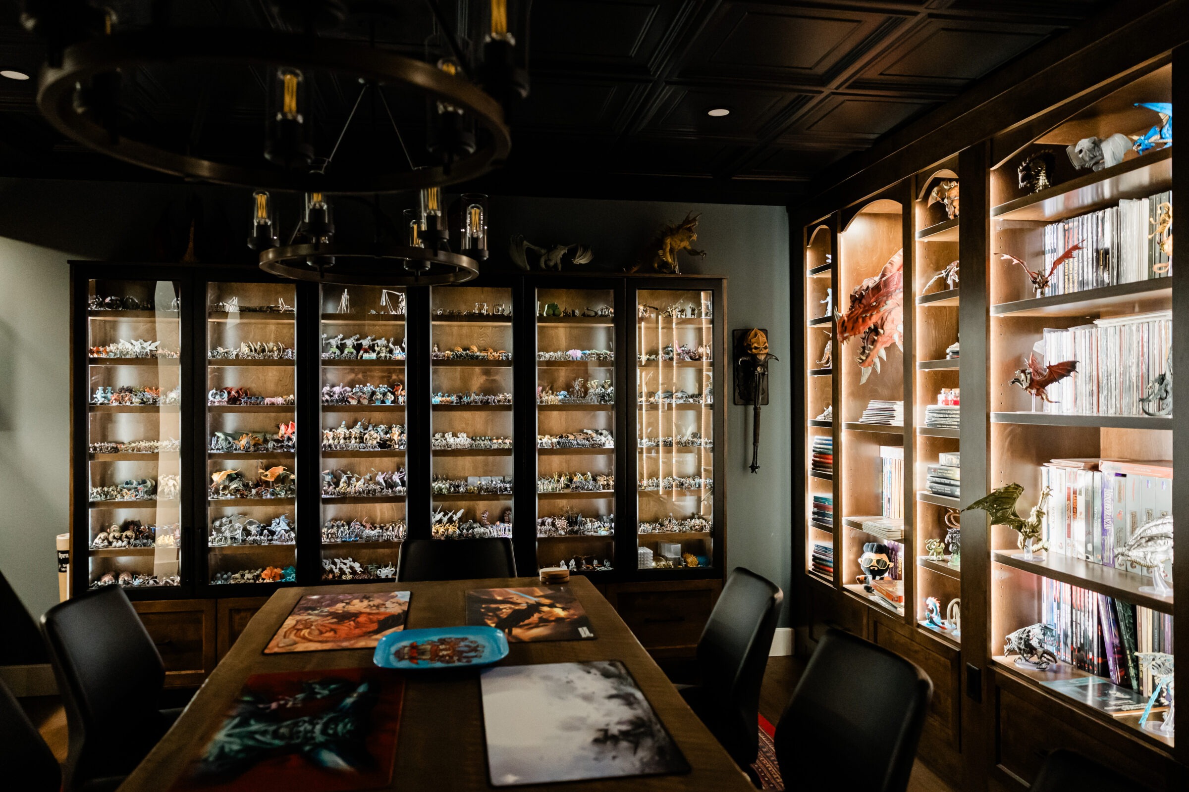 A dimly-lit room with a wooden table, surrounded by chairs, and shelves filled with miniature figures, with additional books and unique items.