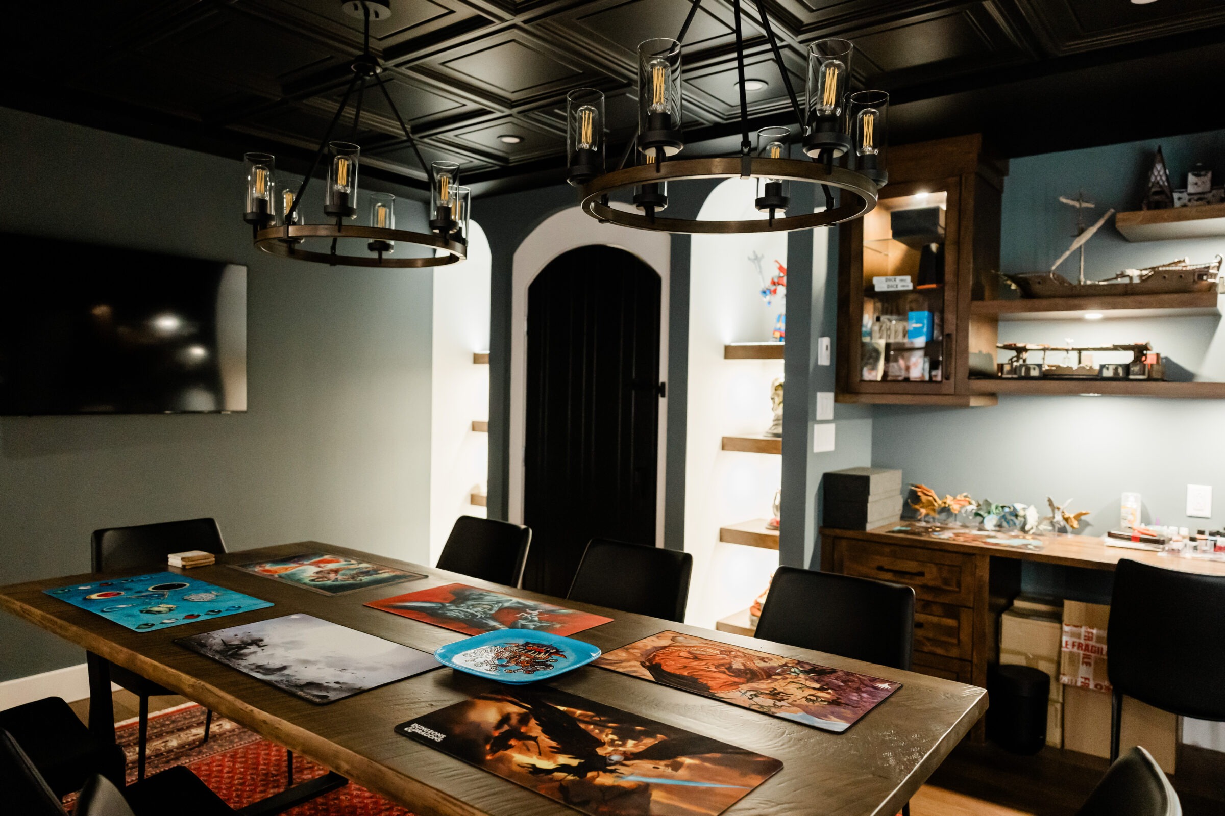 A modern room with a wooden table, art prints, circular chandeliers, and shelves with figurines. A dark palette dominates, with a cozy atmosphere.