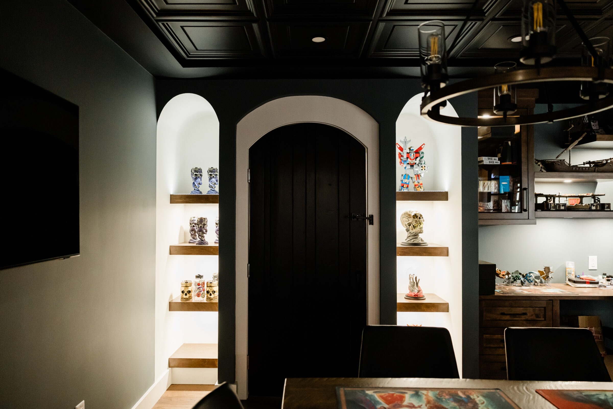 A stylish interior with a black arched door, illuminated alcoves displaying colorful vases, a dark paneled ceiling, and a modern chandelier overhead.